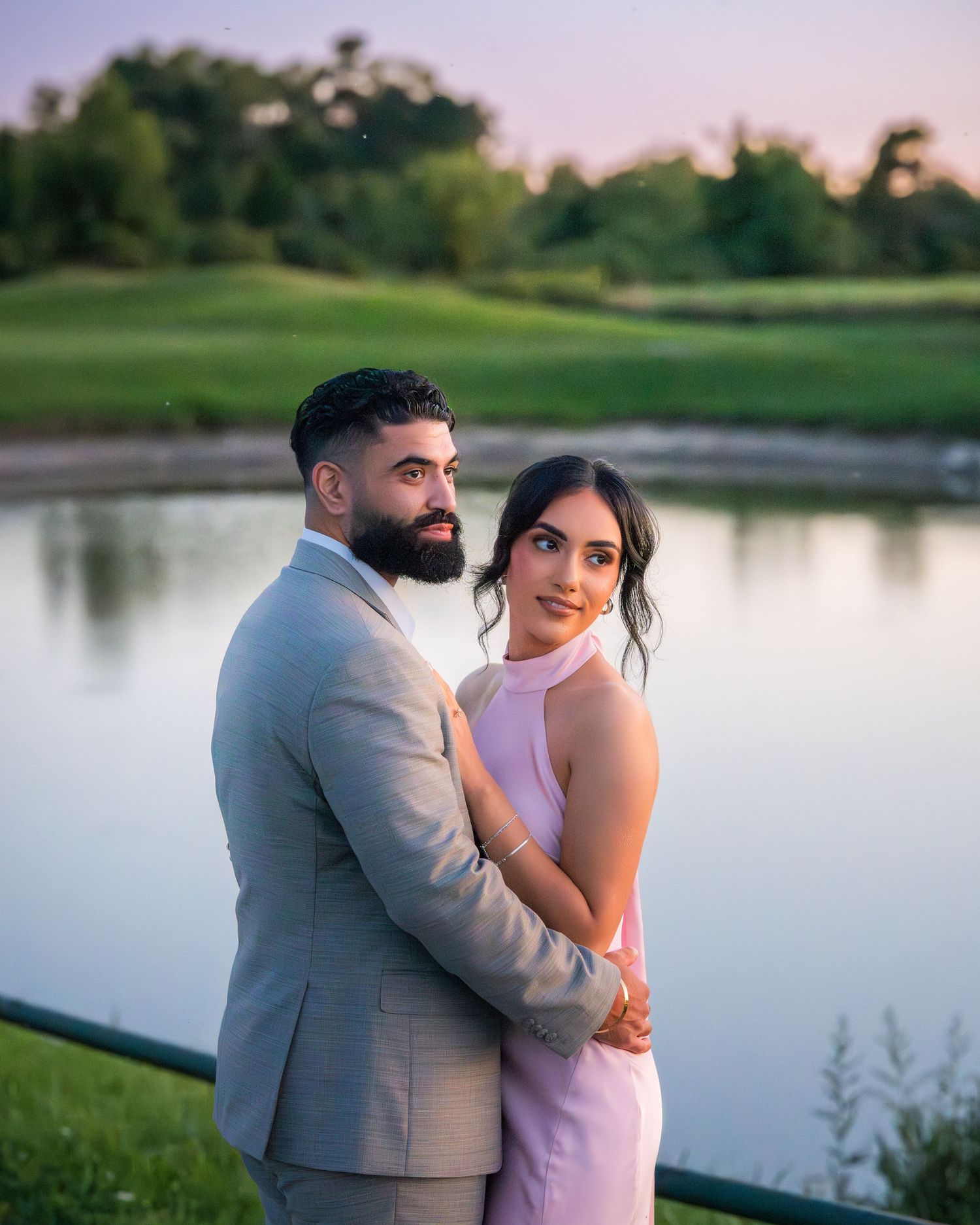 Couple embraces by tranquil lake during sunset photoshoot in formal attire.