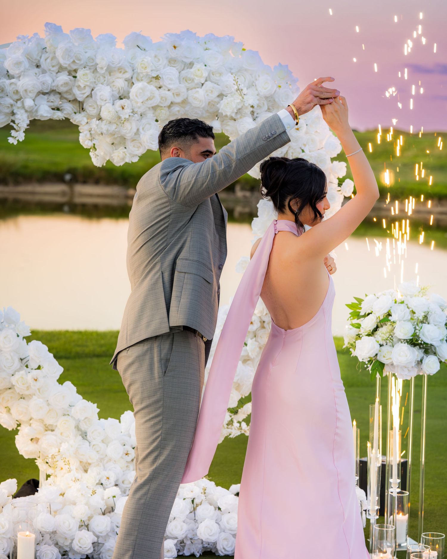 A couple dances beneath a white floral arch during a romantic sunset celebration.