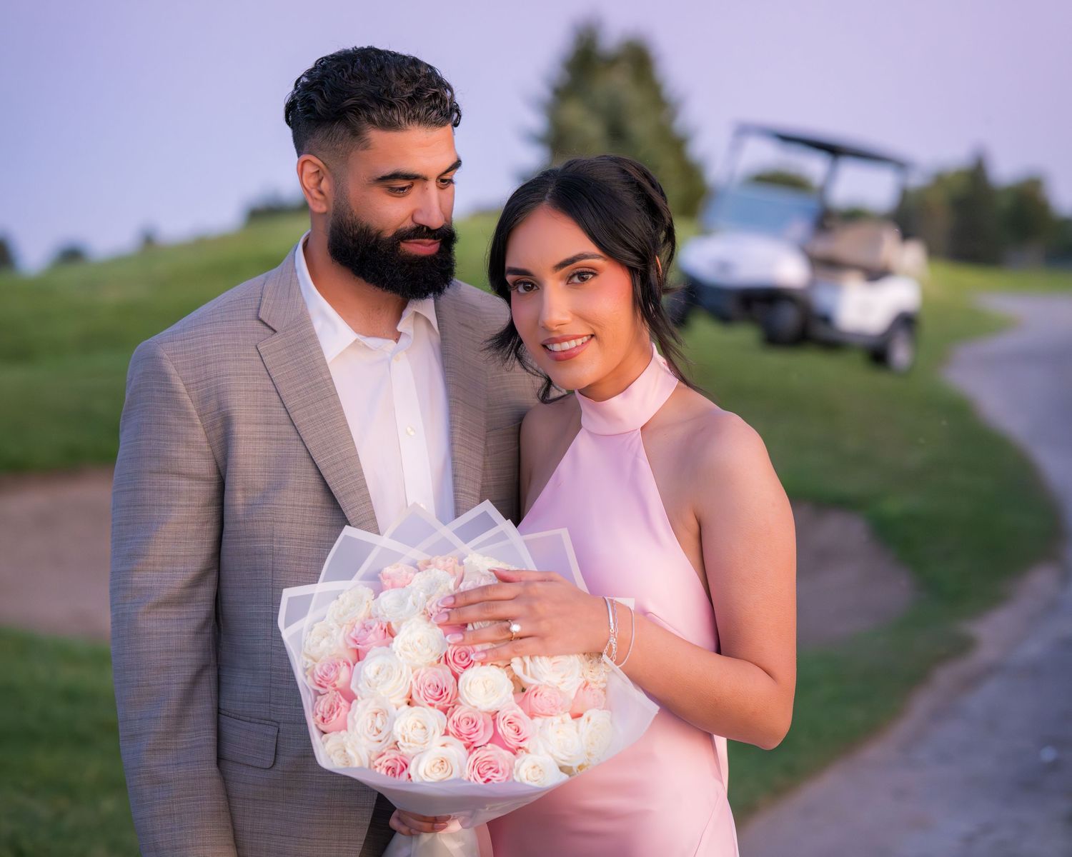 A couple in pink and grey formal wear walks together holding a bouquet of pink flowers.