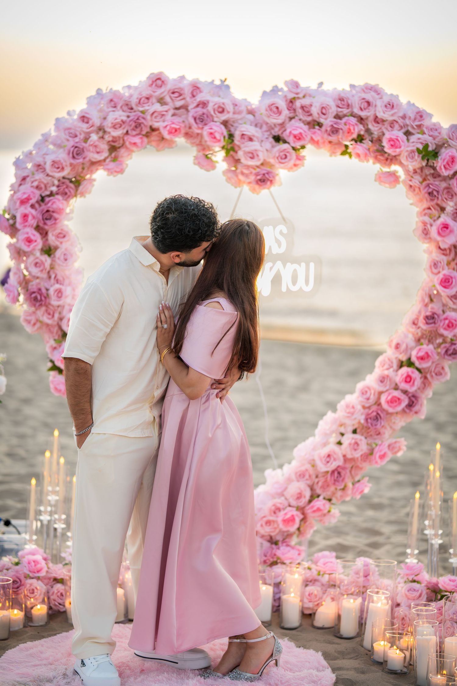 Intimate beach proposal scene with pink floral heart arch, candles, and couple in white and pink attire.
