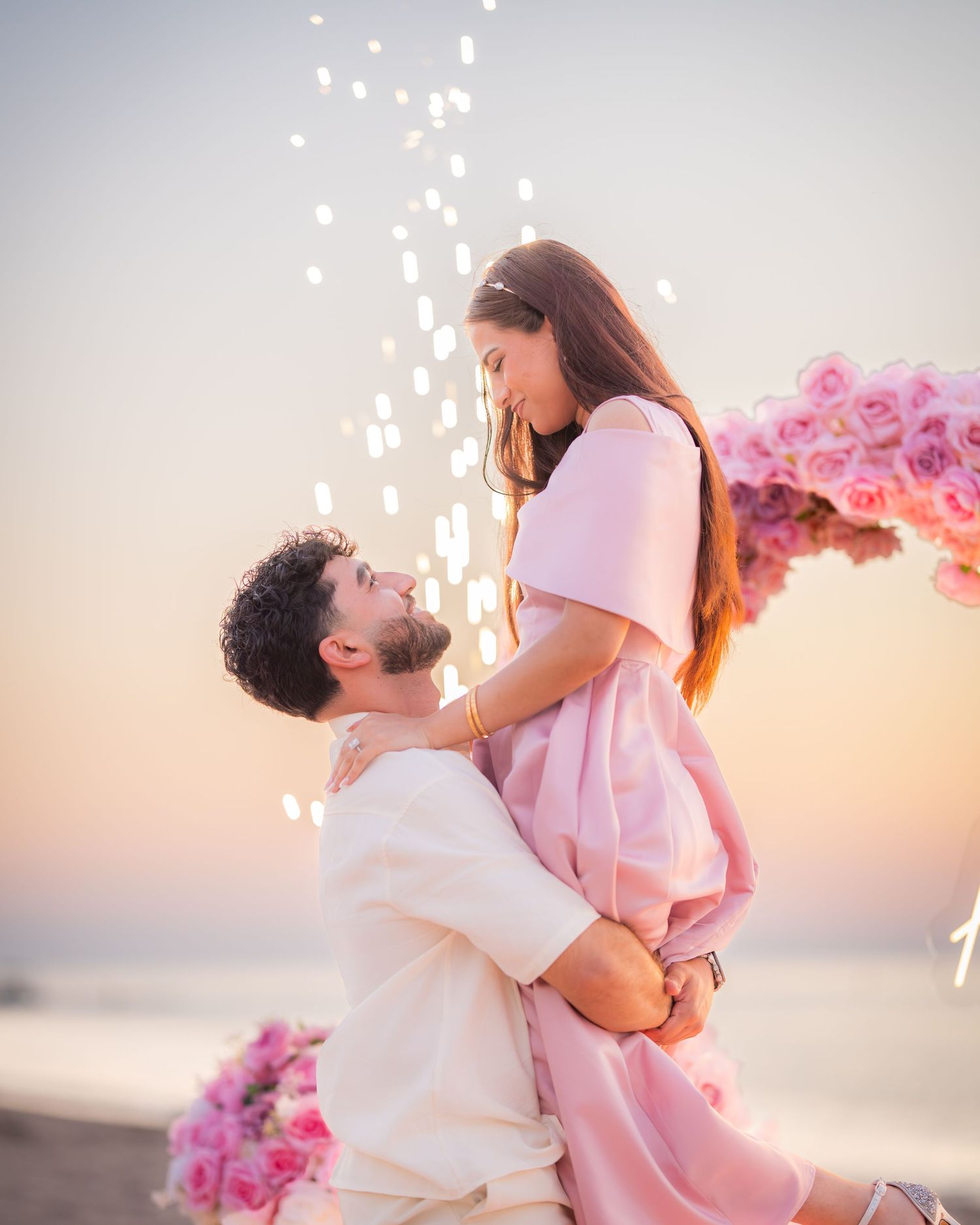 Couple celebrating with sparklers under pink floral heart arch on beach.