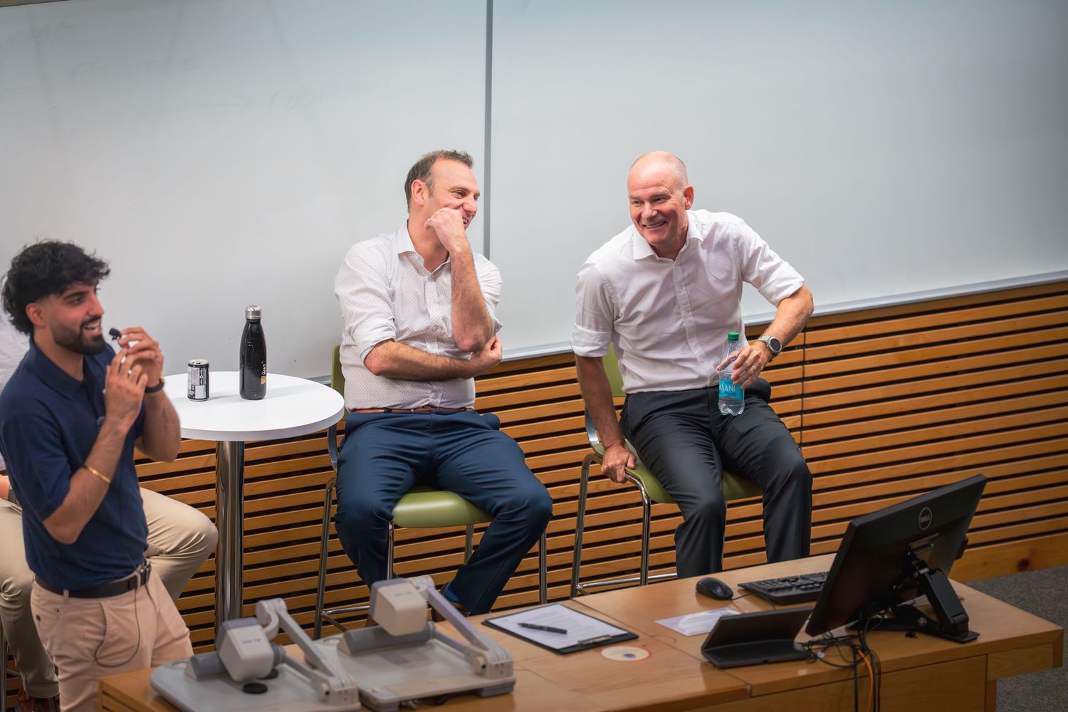 Speakers engage in animated conversation while seated on wooden benches against white wall.