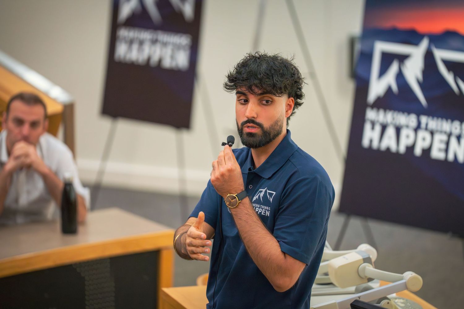 A presenter in a navy blue polo shirt gestures while speaking to an audience in a lecture hall.
