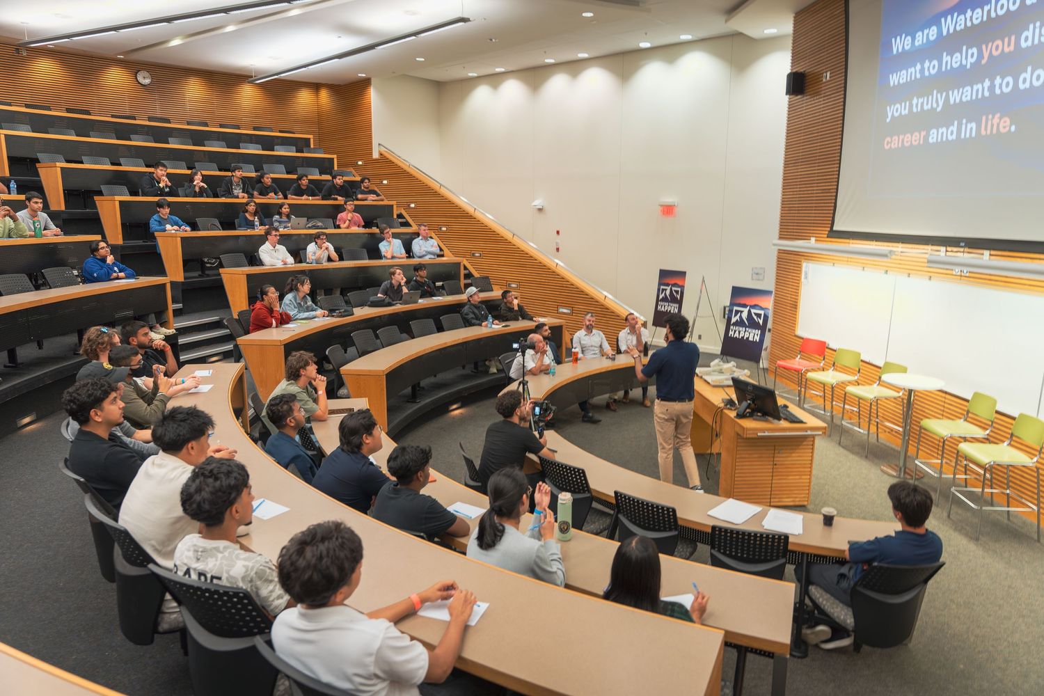 Students sit in tiered rows of a modern lecture hall during an academic presentation.
