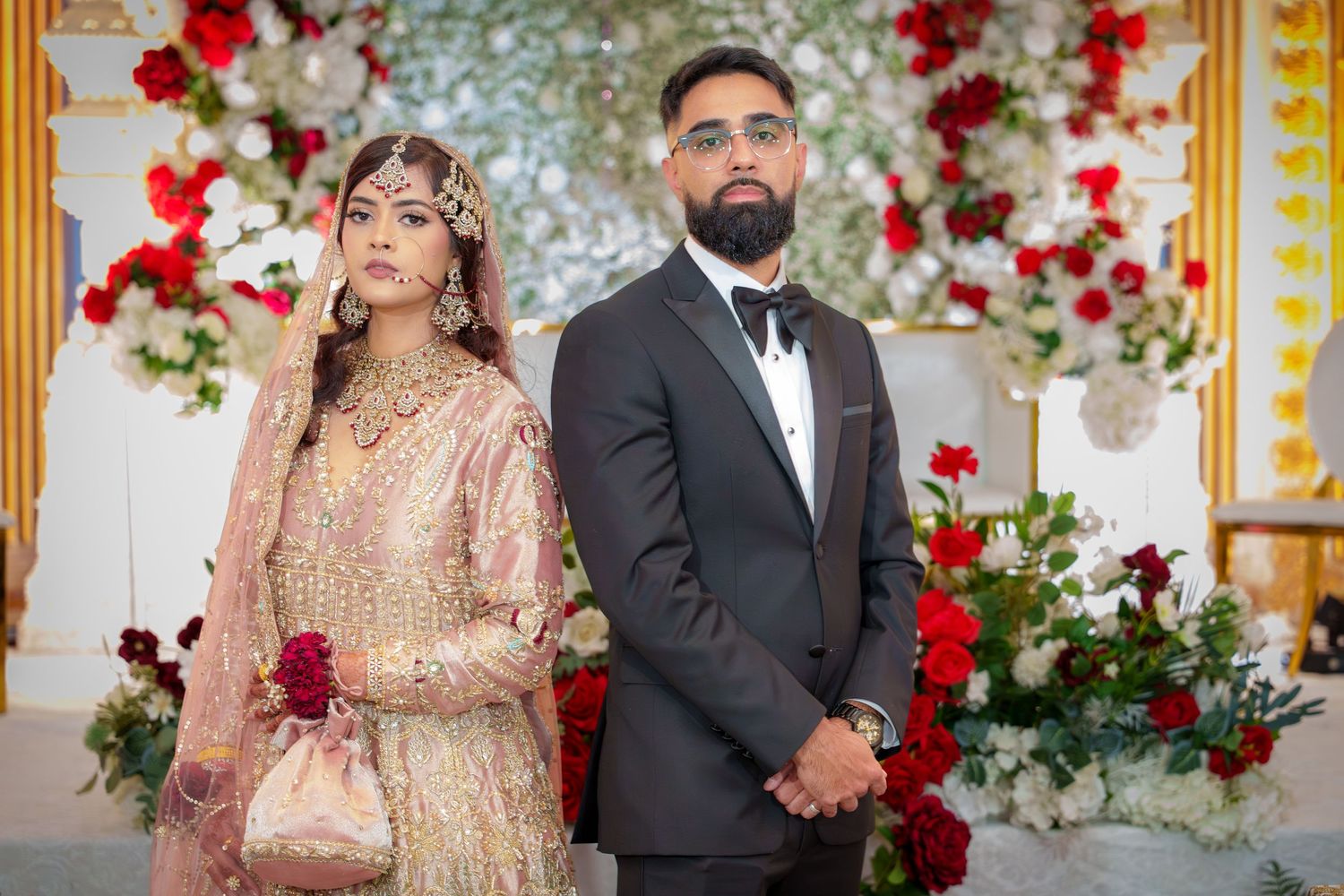 South Asian wedding couple poses against floral backdrop with red and white roses.