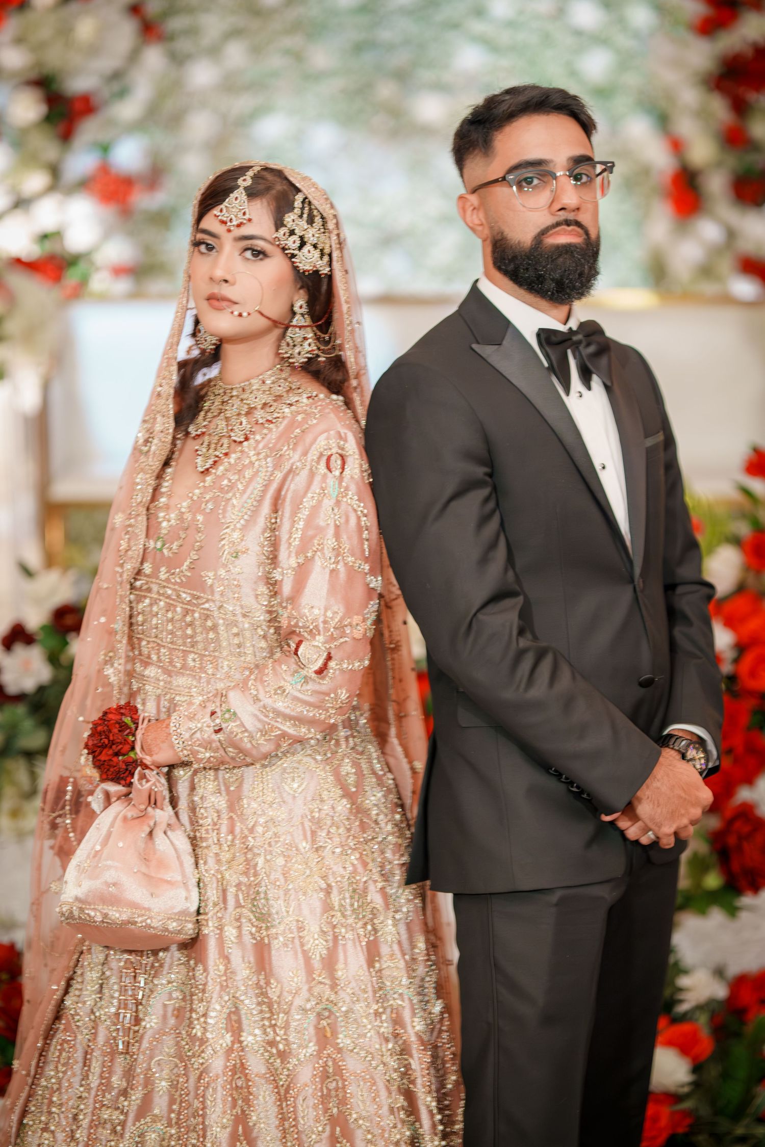 Traditional South Asian wedding couple in peach lehenga and black tuxedo against floral backdrop.