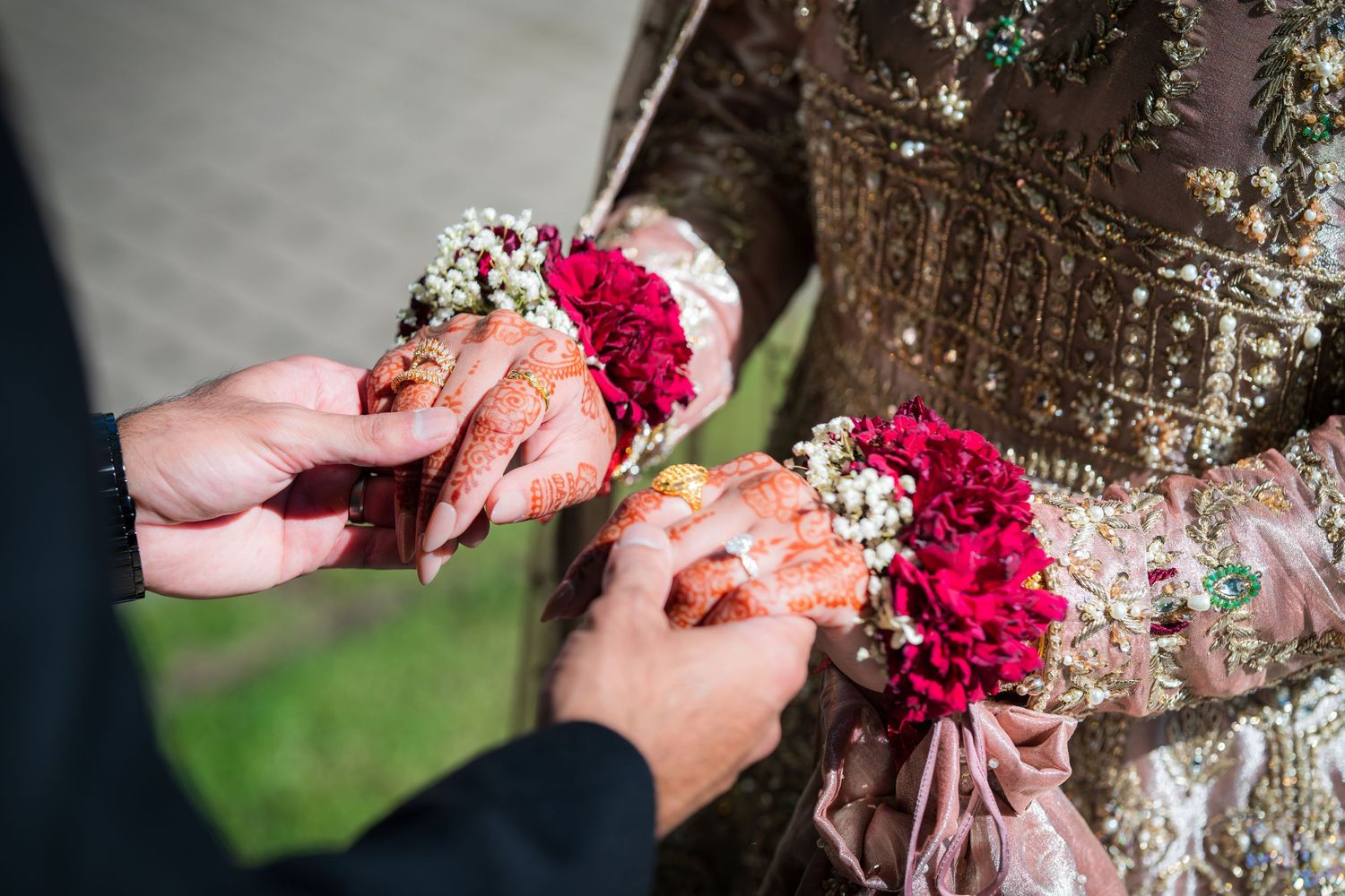 Couple's hands placing wedding rings during an outdoor ceremony with red floral decorations.