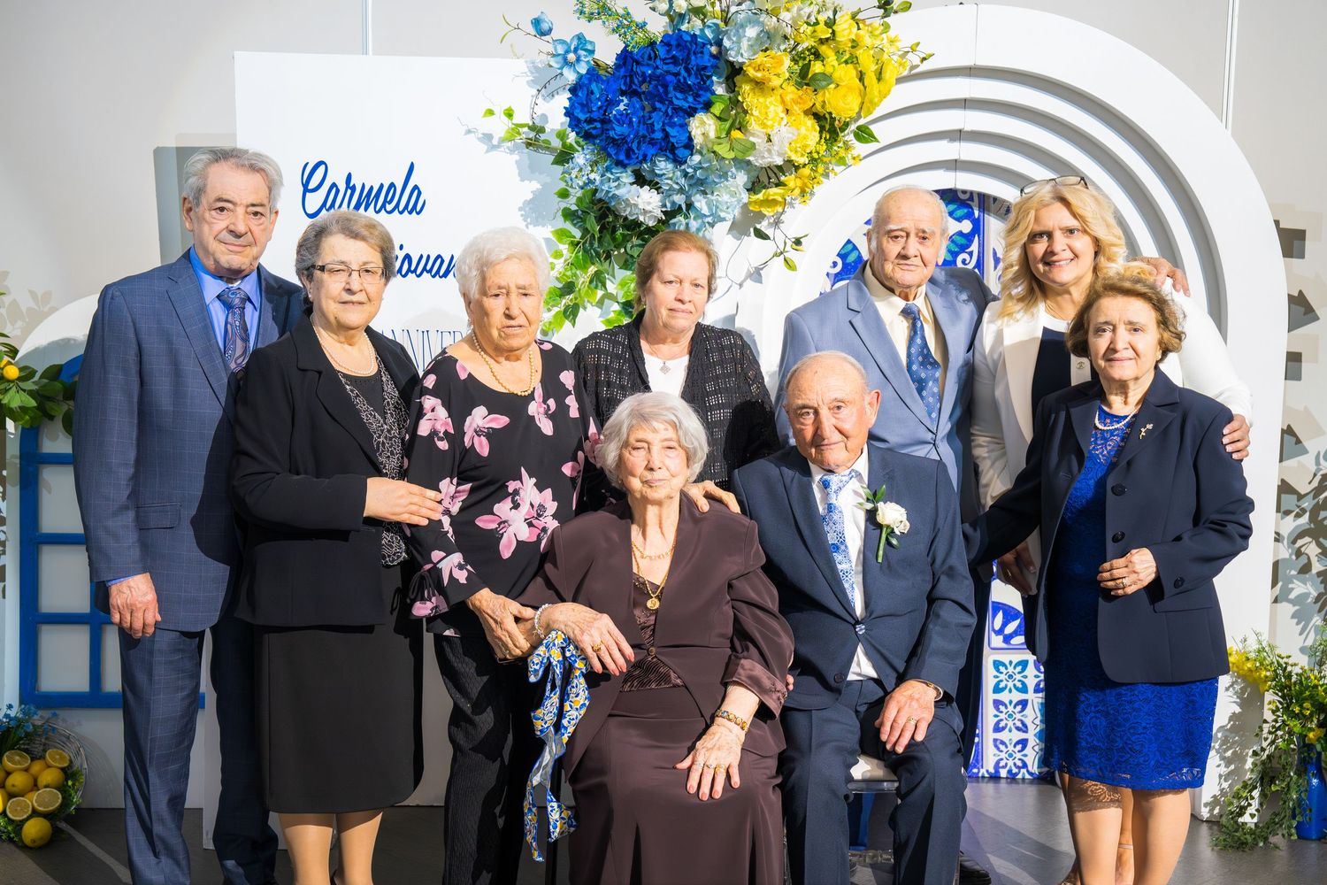 Group portrait of elderly family celebrating a milestone anniversary under a floral arch with blue and yellow flowers.
