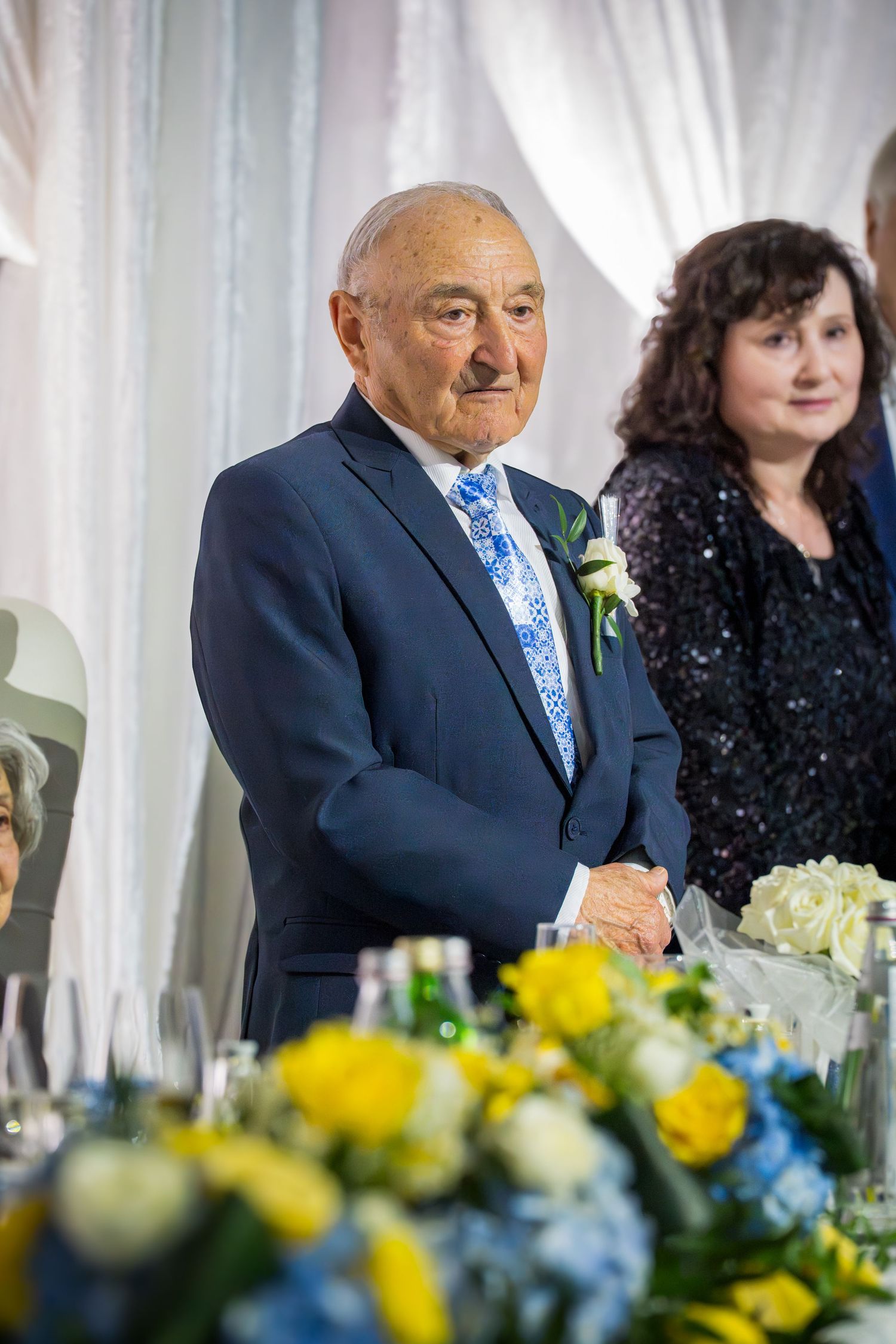 Senior man in navy blue suit standing at wedding reception head table with floral arrangements.