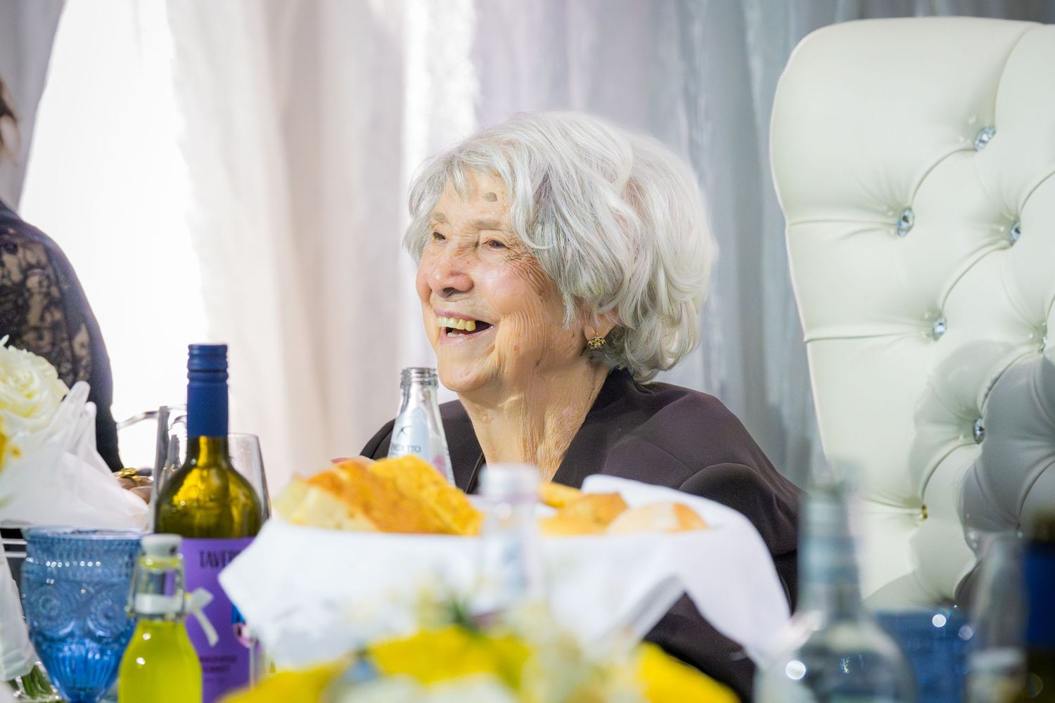 Smiling elderly woman with white hair seated at a decorated celebration table with blue glasses and yellow flowers.