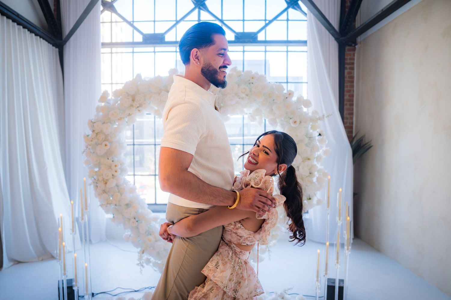 Happy couple embracing and smiling in front of large heart-shaped white floral arrangement.