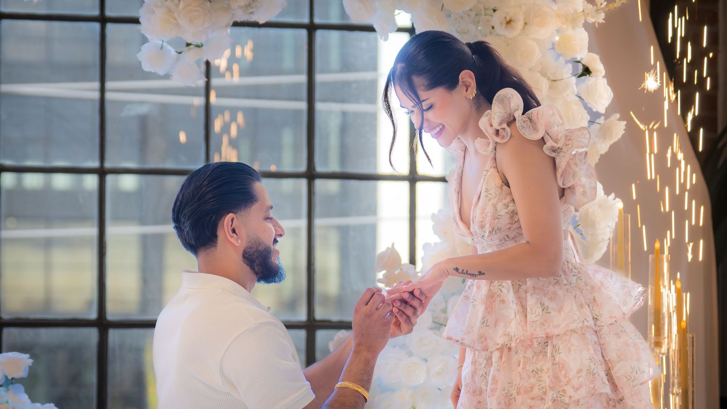 Romantic marriage proposal with man on one knee before white floral arch backdrop.