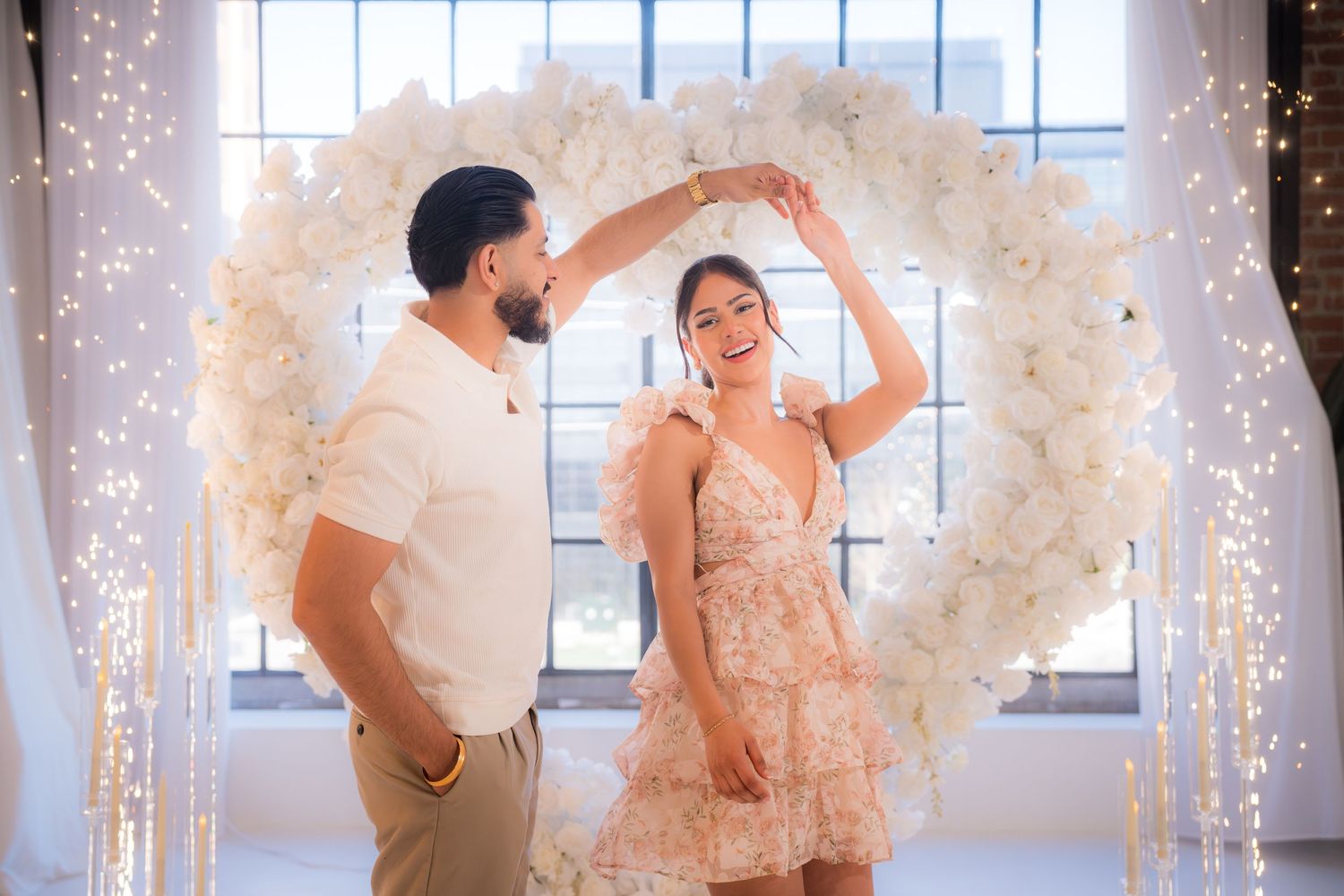 Man spinning woman in floral dress under white heart flower arch with fairy lights.