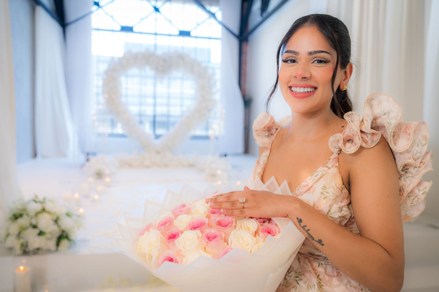 Smiling bride holding bouquet of pink roses in front of white heart-shaped floral backdrop.