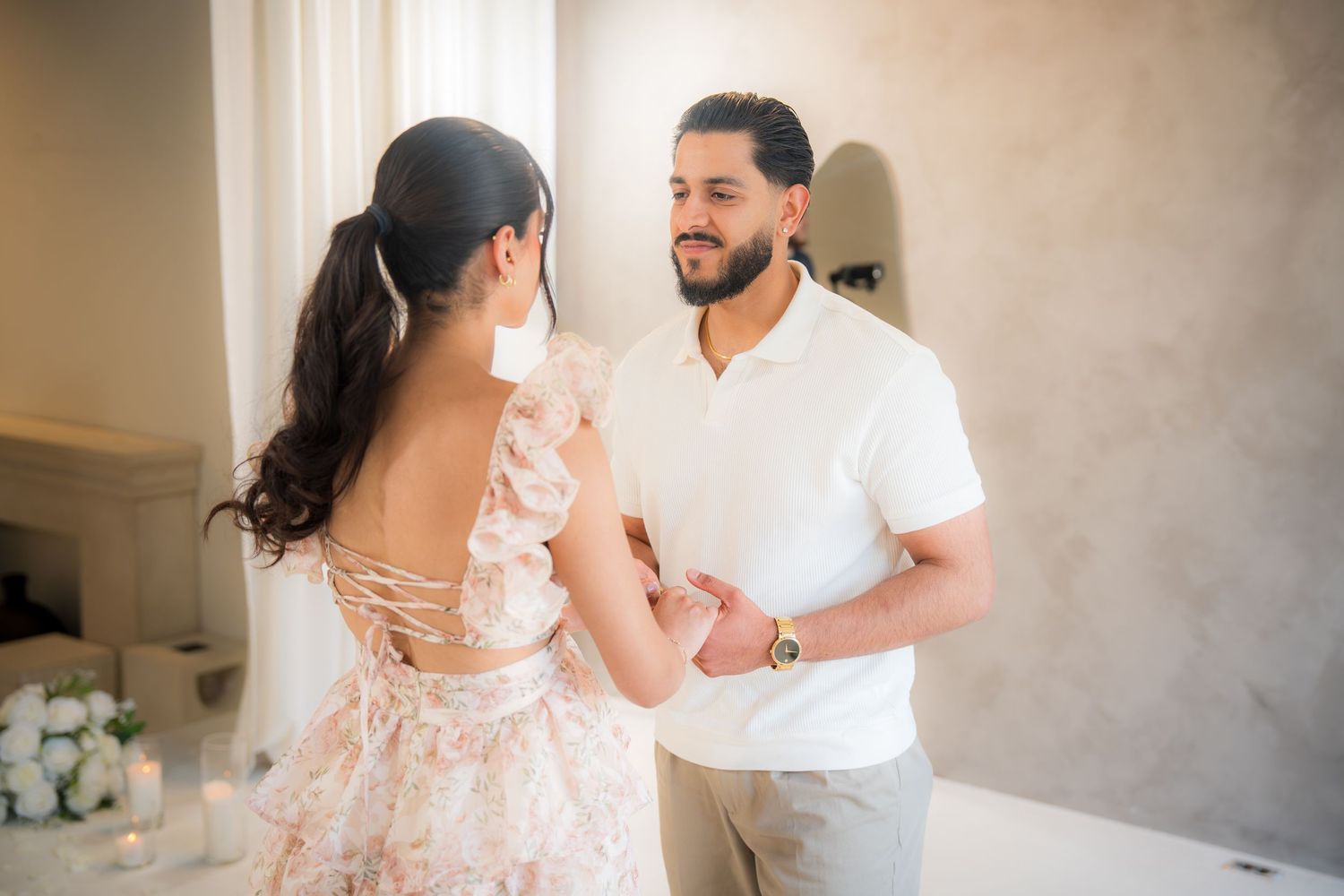 Couple holding hands and smiling at each other indoors, woman wearing a floral backless dress during a romantic moment.