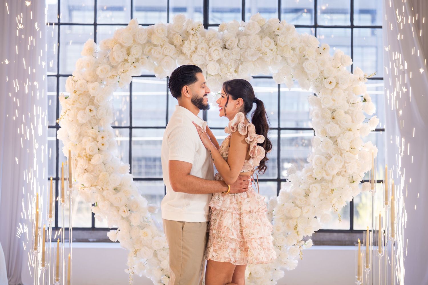 Couple embracing and gazing at each other in front of large white floral heart arch with fairy lights.
