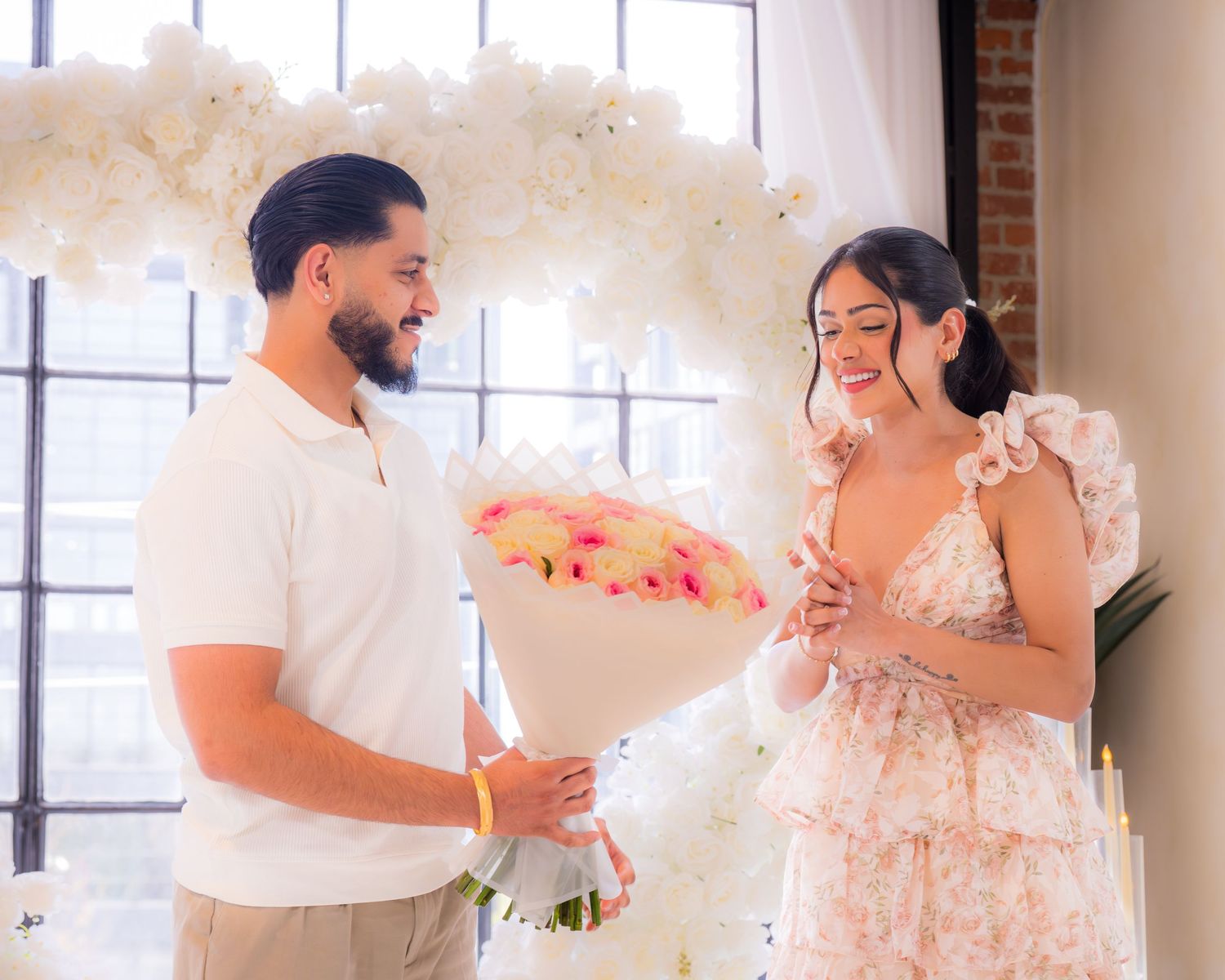 Man presenting a bouquet of pink roses to a smiling woman in a floral dress at a proposal setup.
