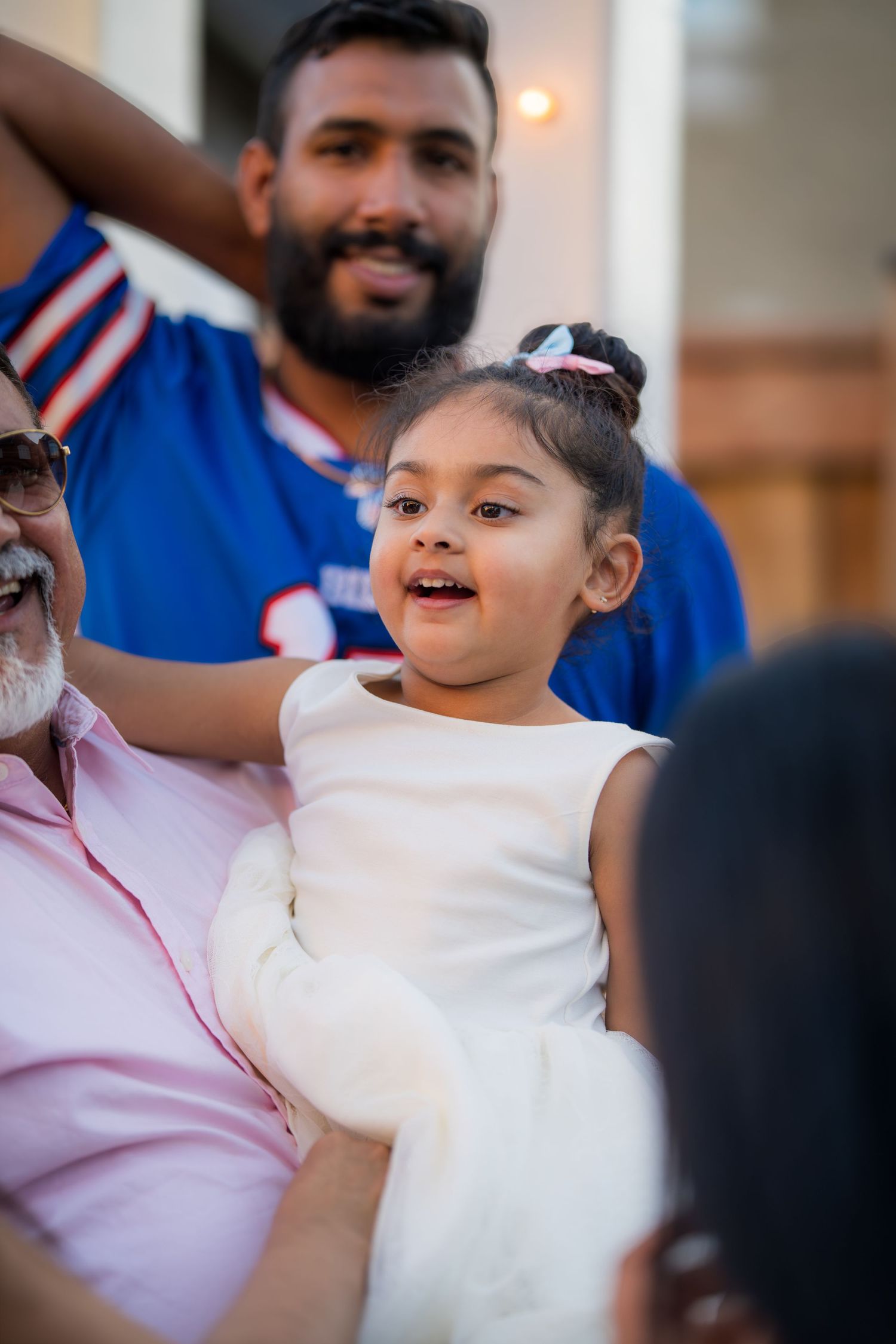 A cheerful toddler girl in white dress is held by an older man while a bearded man smiles behind them.