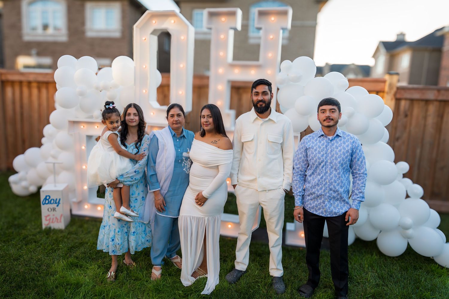 Family group poses in front of illuminated marquee letters and white balloons at a gender reveal.