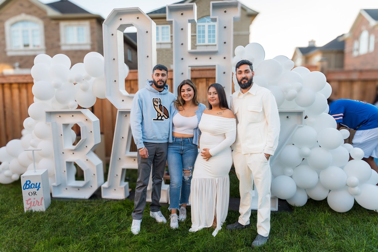 Group of four friends smiling at an outdoor baby shower with white balloon arch and lit letter decor.