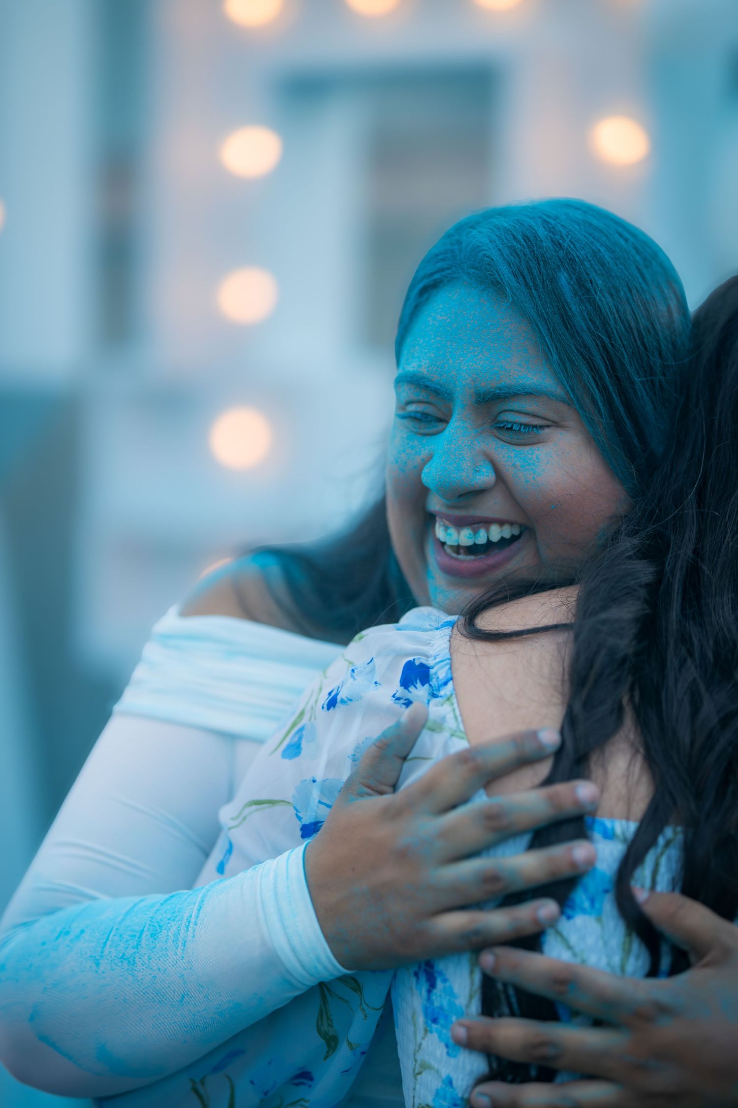 Two women with blue Holi powder on their faces share a joyful hug while laughing together.