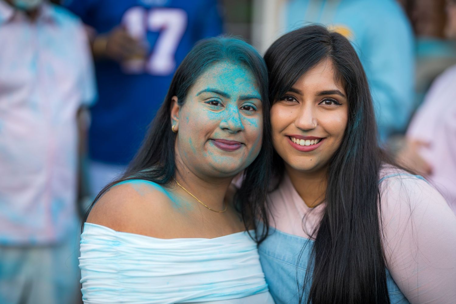 Two women smiling together covered in blue gender reveal powder at an outdoor celebration event.