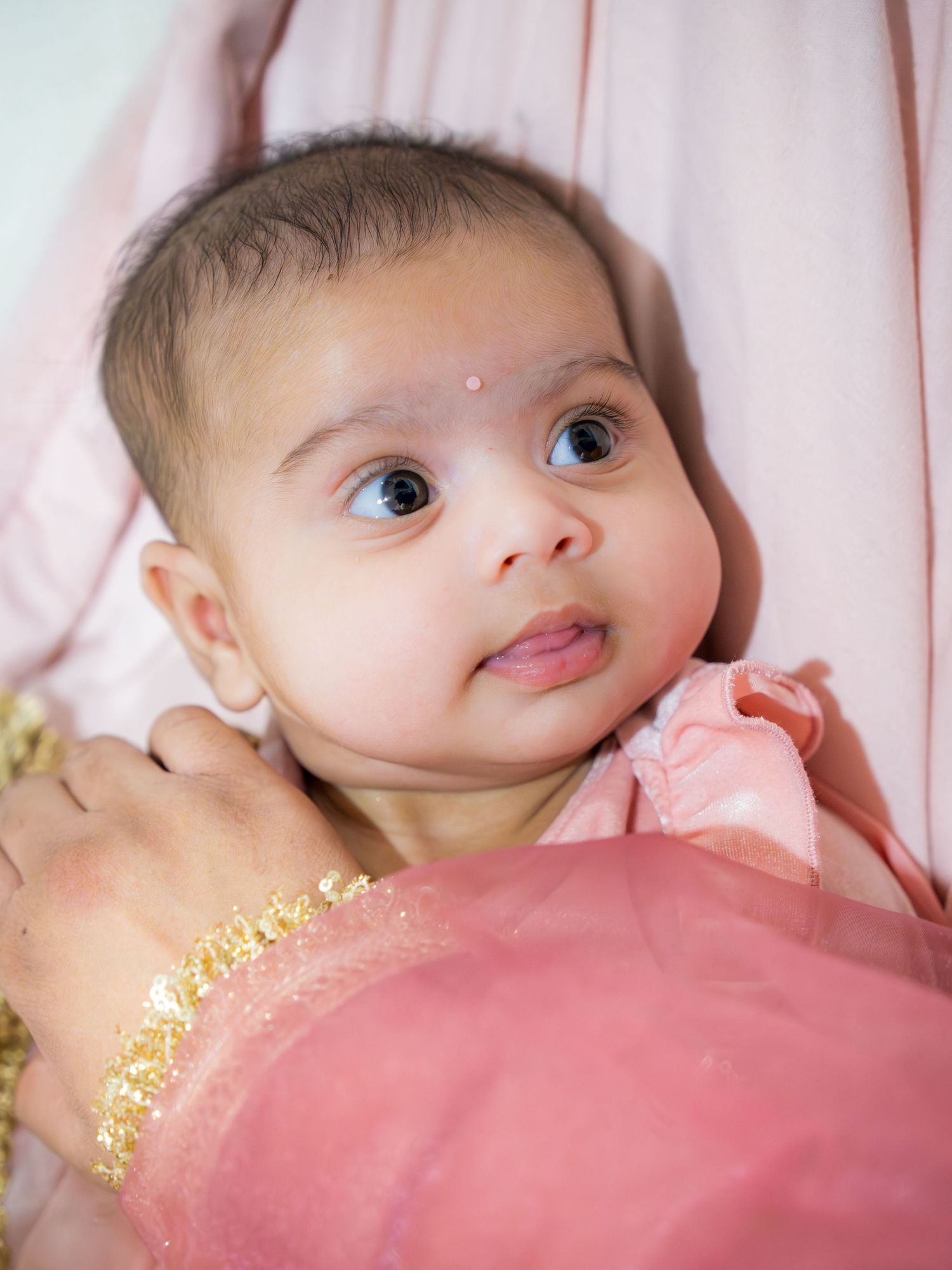 Adorable baby girl in pink outfit with gold jewelry and red bindi dot at Indian Lohri celebration.
