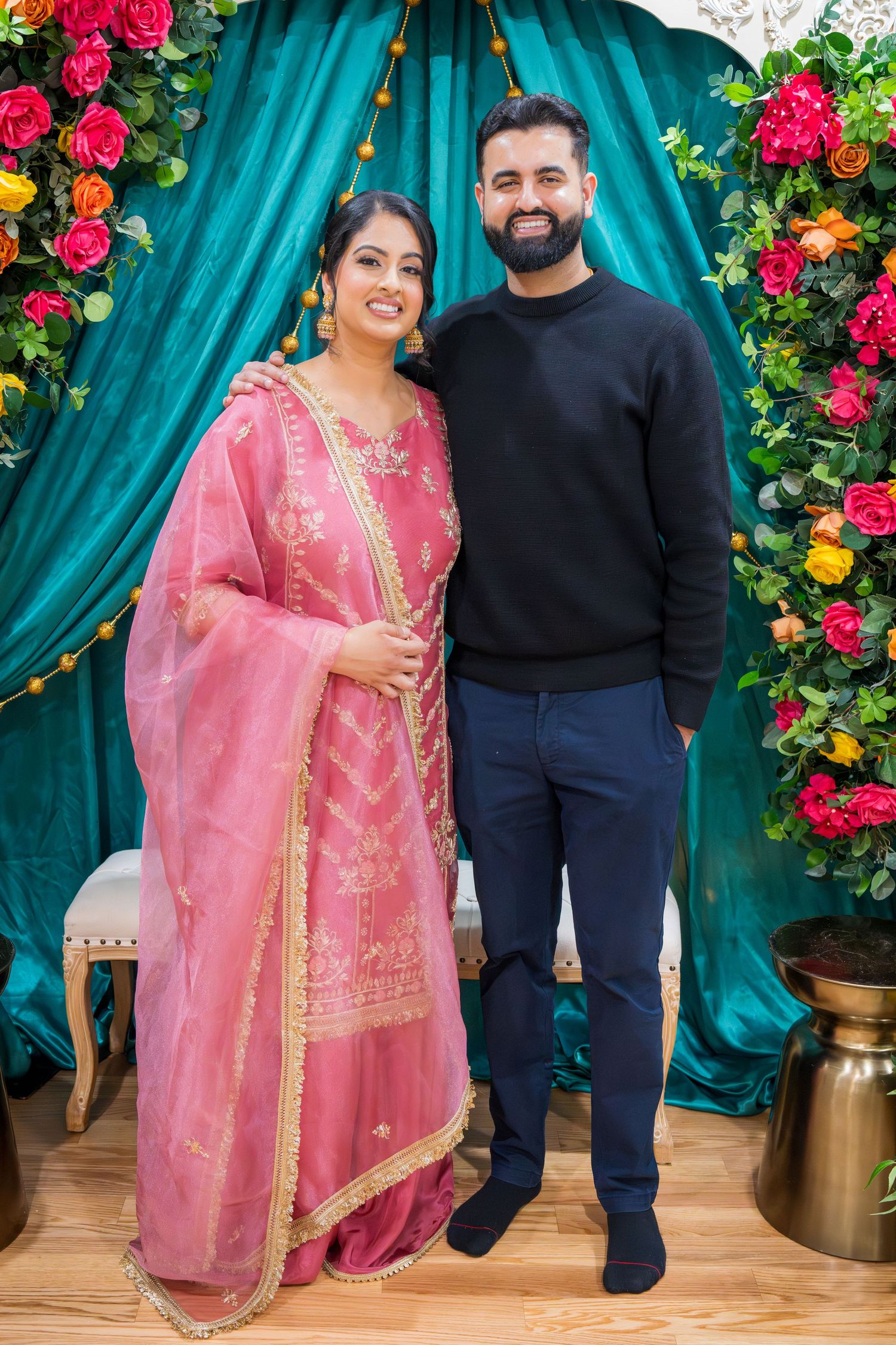 Woman in pink salwar kameez and man in black casually posing at a floral South Asian event.
