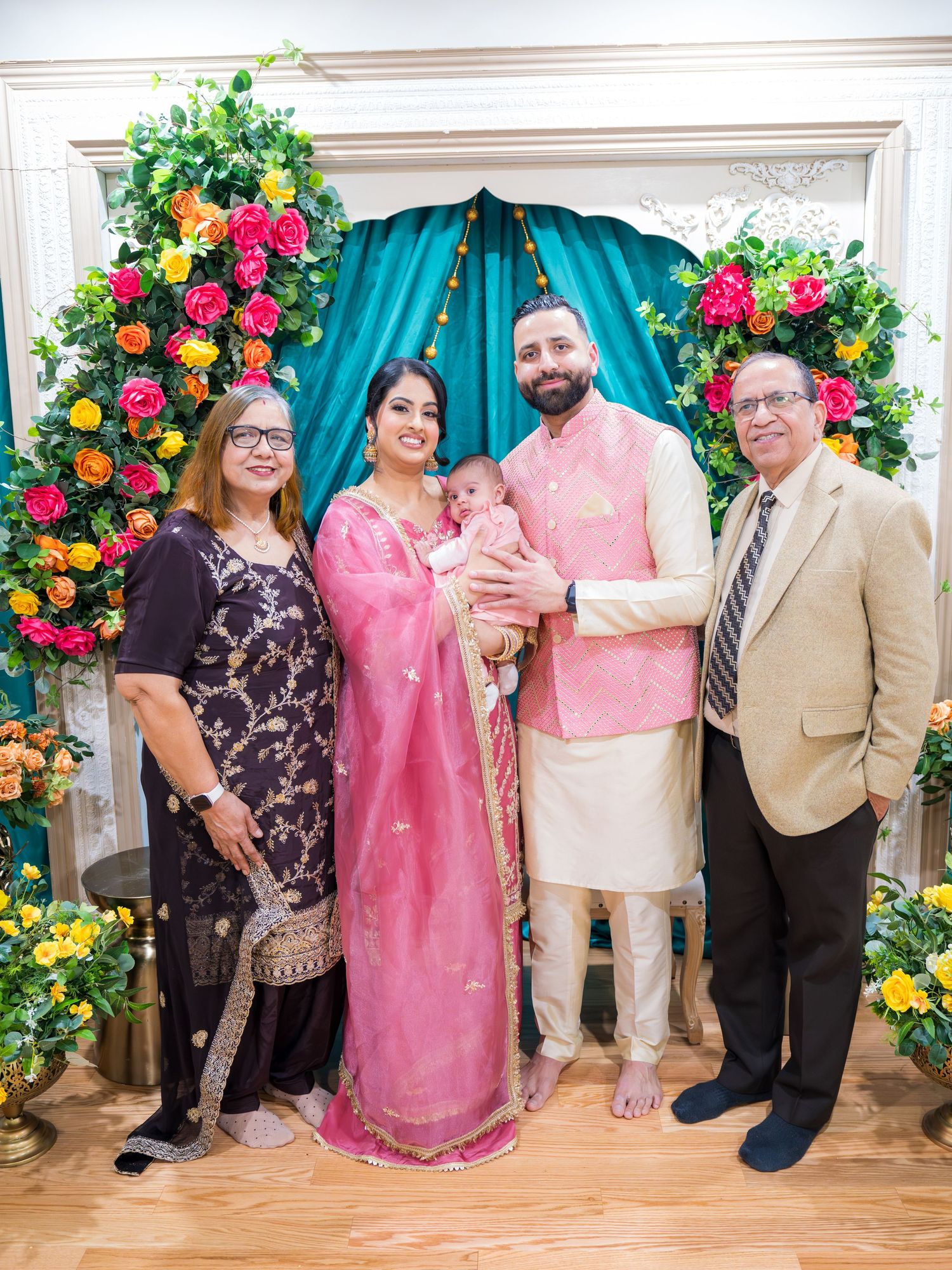 A family of four in formal attire poses with a newborn baby in front of a colorful floral arch backdrop.
