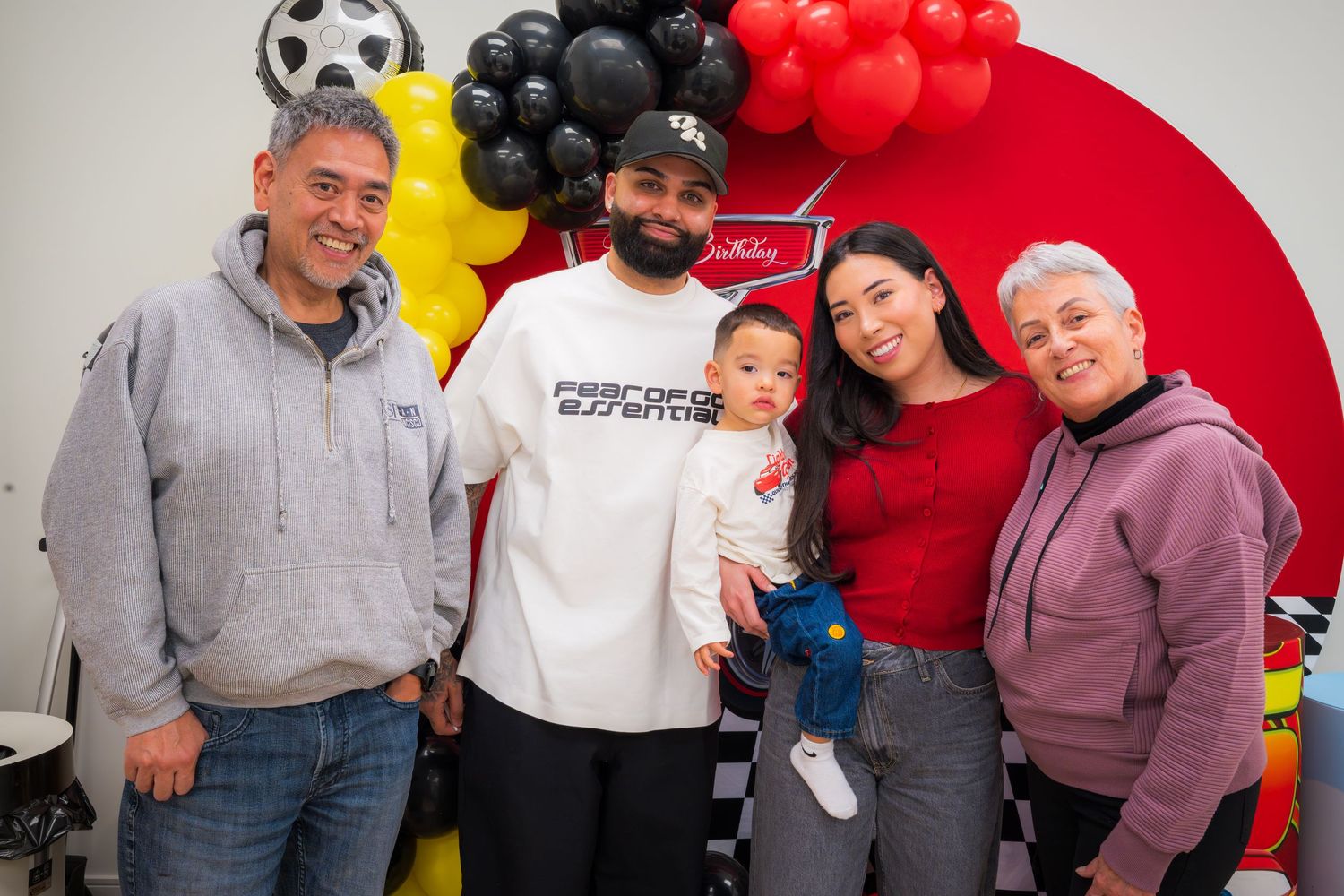 A family of five poses together at a Cars-themed first birthday party with colorful balloons.