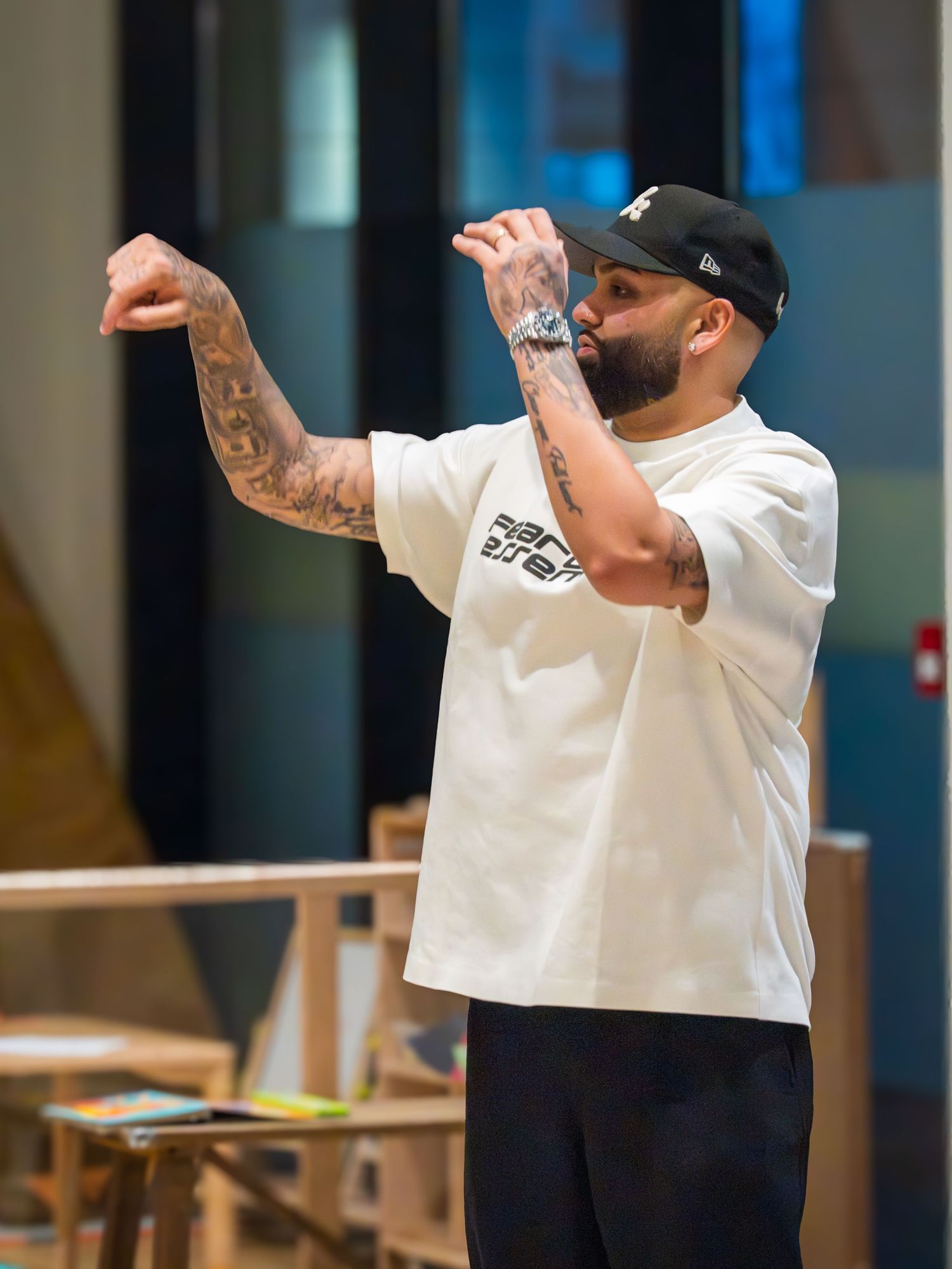 A tattooed man in a white t-shirt and black cap gestures with his hands in an indoor setting.