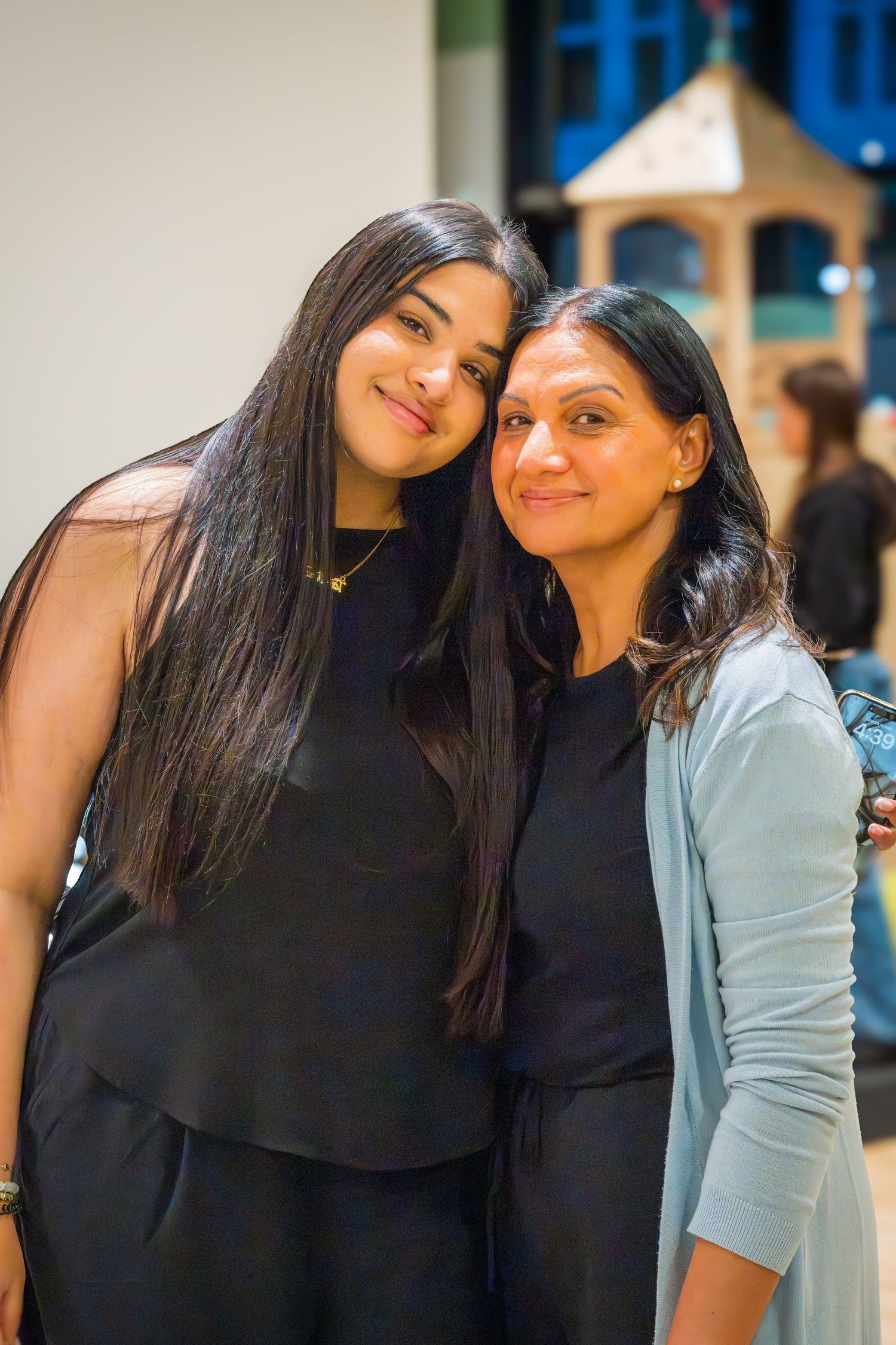 Two women with long dark hair smiling together at an indoor event, one wearing a light blue cardigan.
