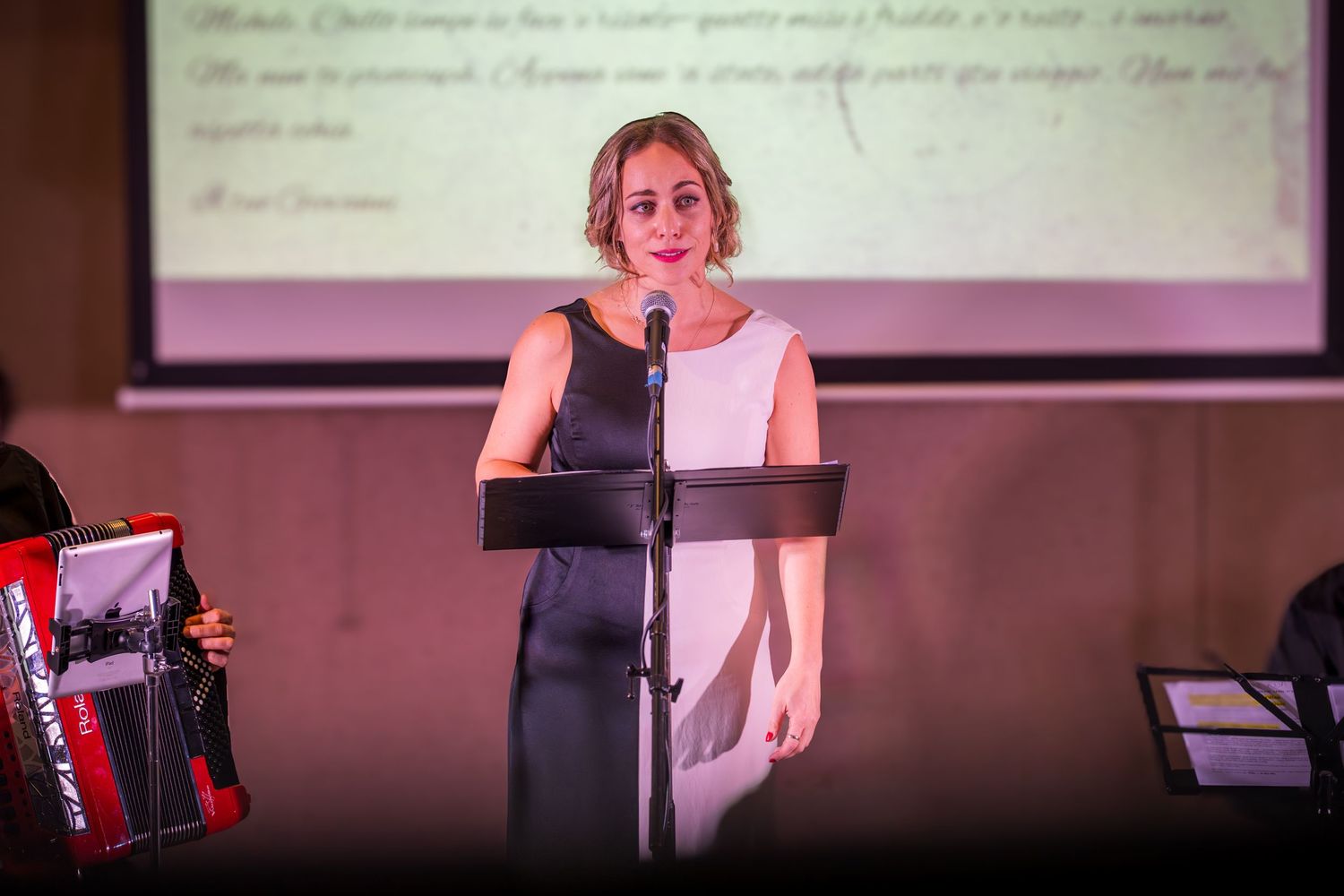 Woman in elegant dress speaking at podium during a live musical performance event on stage.