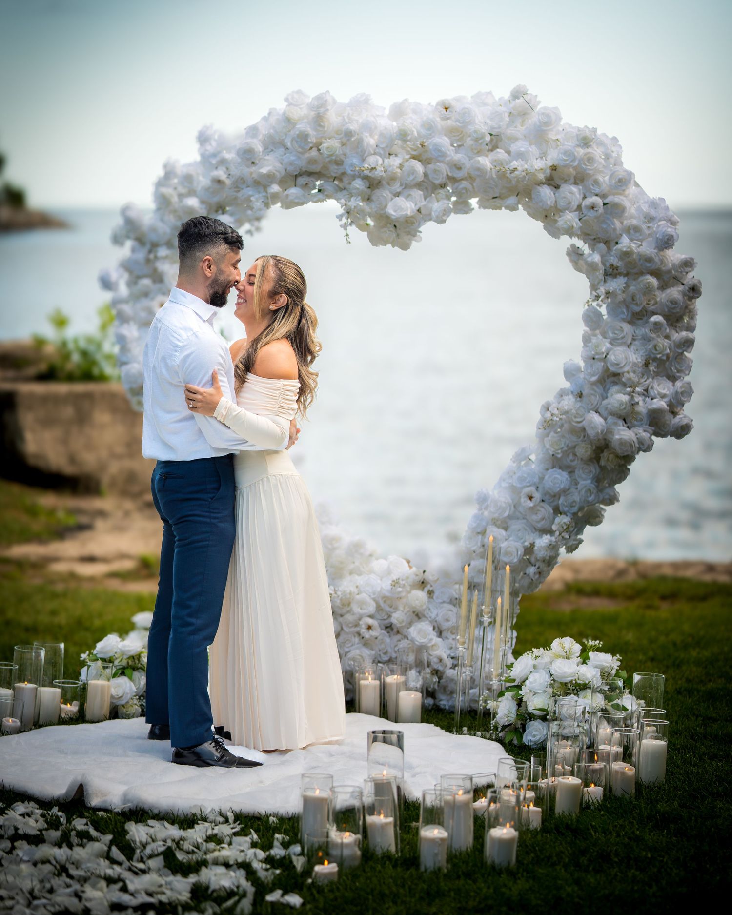 A romantic seaside proposal setup with heart-shaped balloon arch and scattered candles on white platform.