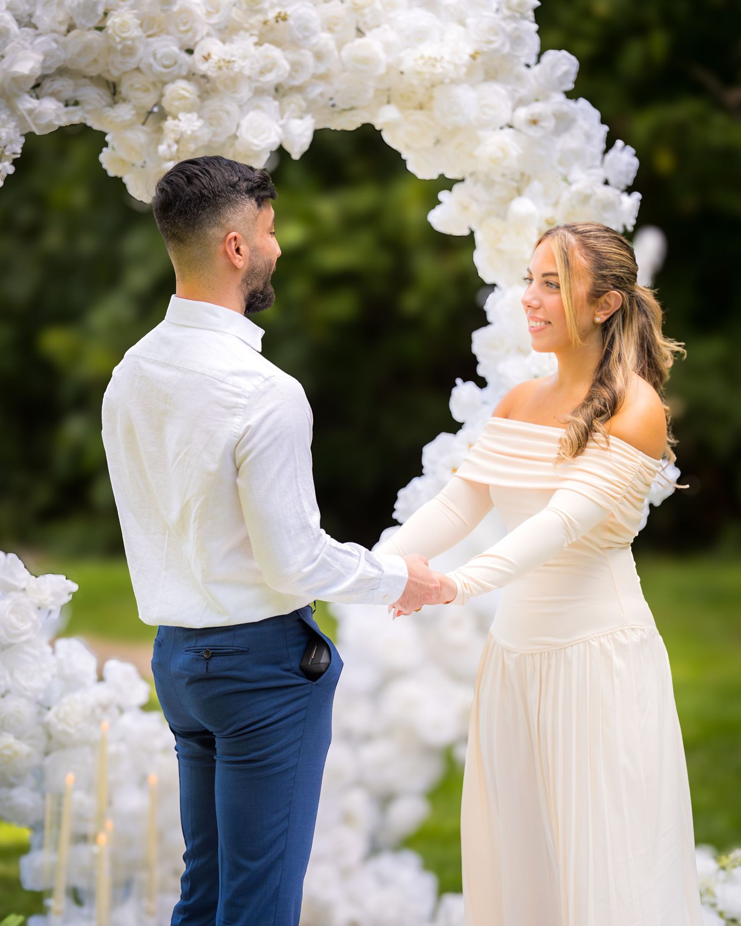Wedding couple shares tender moment under white floral arch during outdoor ceremony.