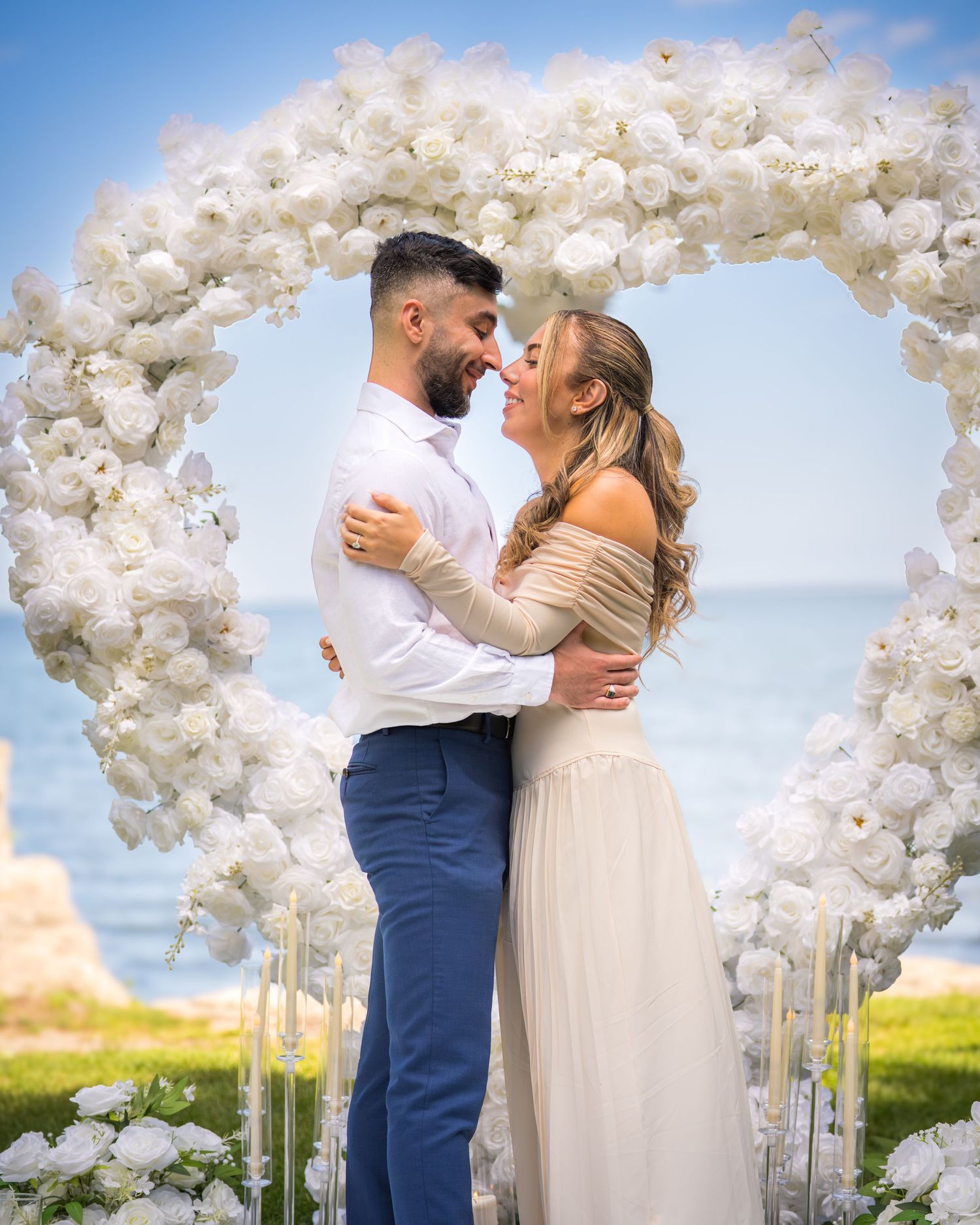 Wedding ceremony setup with white floral arch and candles under blue sky.