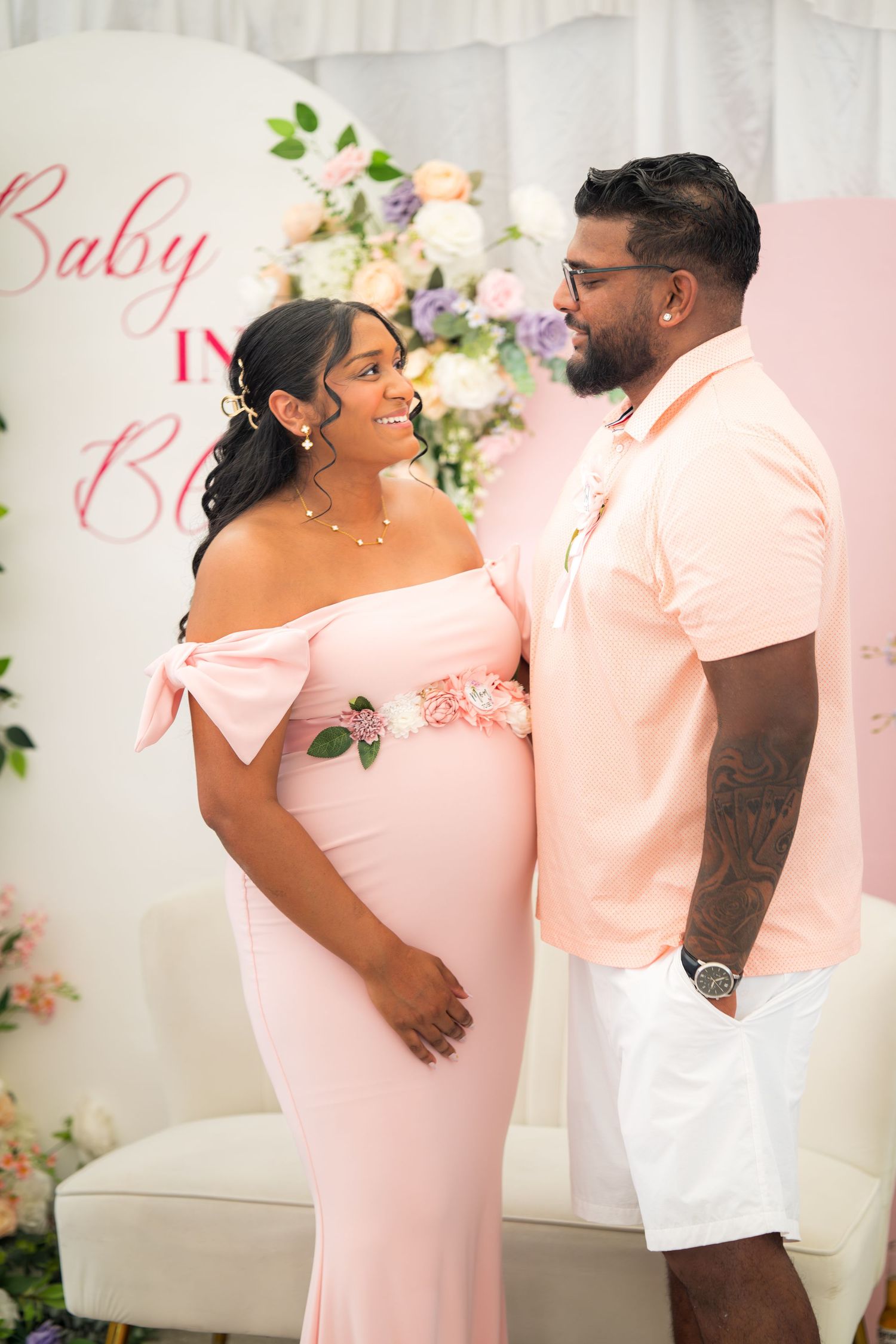 Couple poses at baby shower with floral backdrop and 'Baby in Bloom' text, wearing coordinating pink and white outfits.