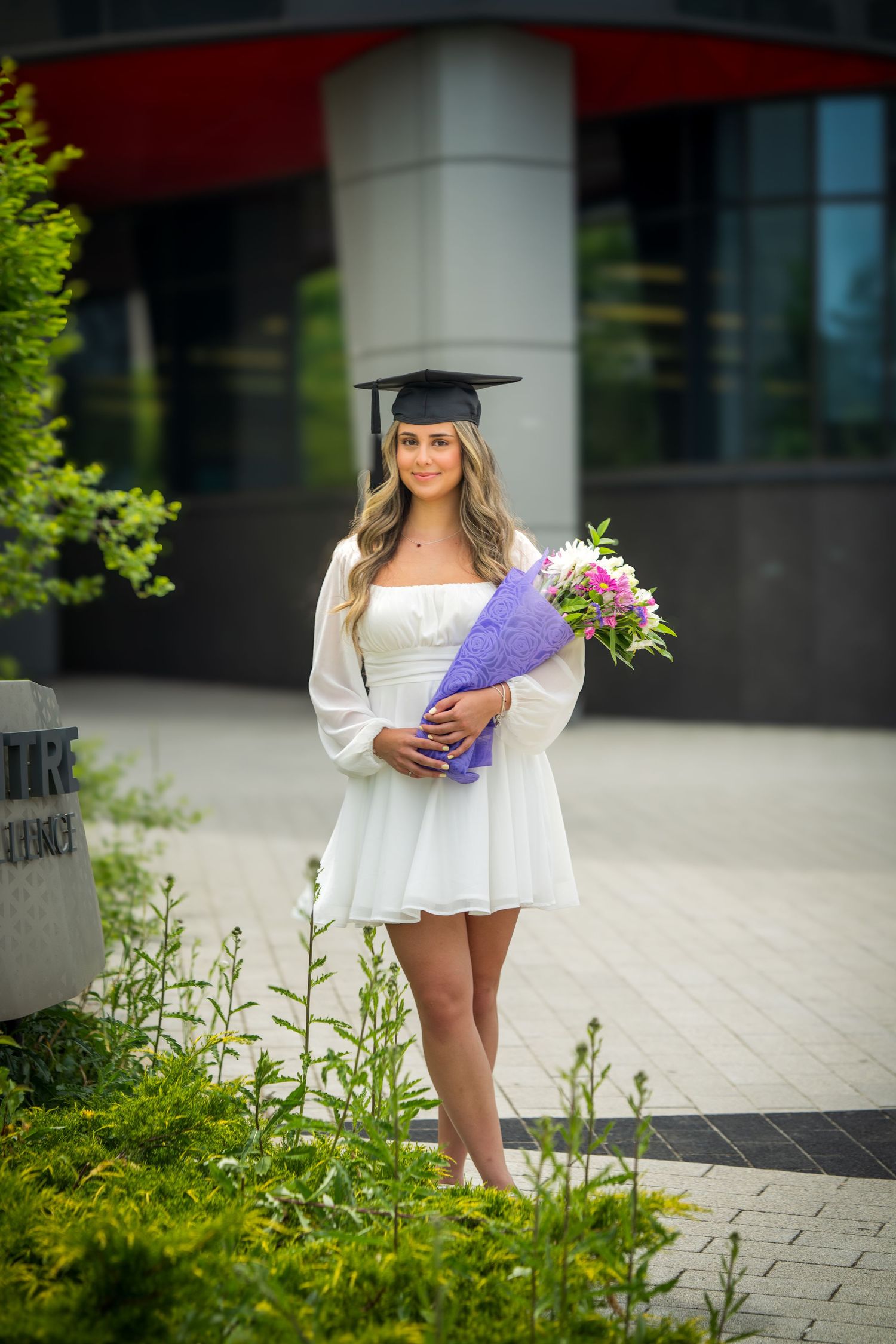 Graduate in flowing white dress holds purple diploma holder near modern building entrance.