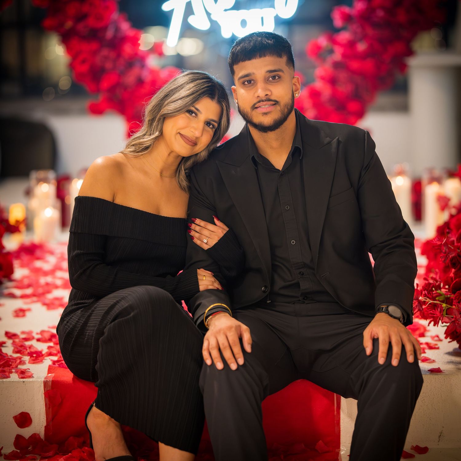 Couple sits together on red surface surrounded by rose petals and neon lighting.