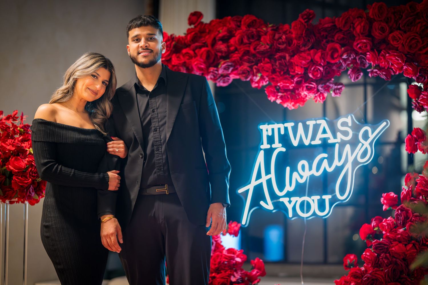 Elegant couple poses by neon blue sign and red rose wall decoration in formal wear.