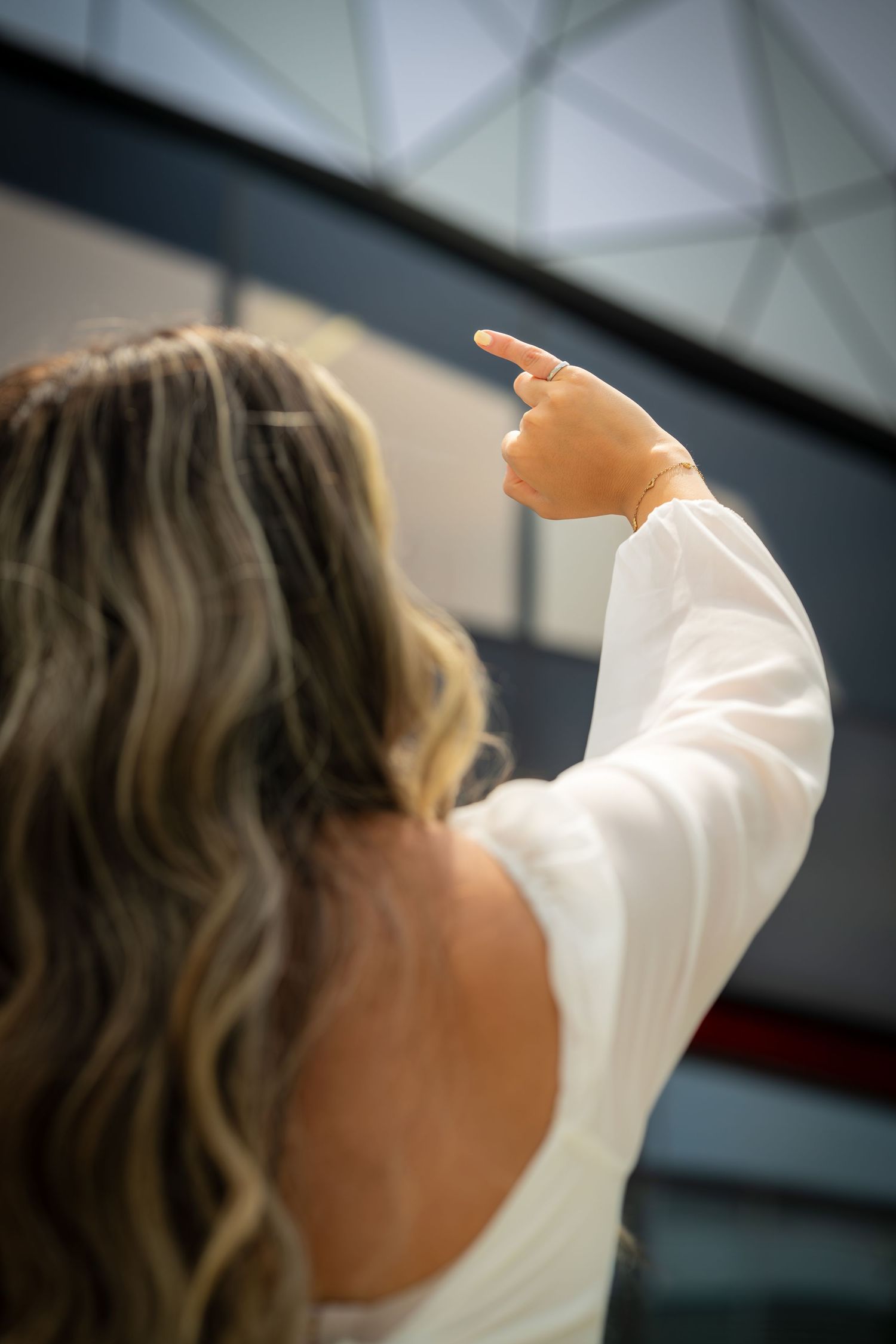 A person with wavy hair in a white sweater points upward while facing away from the camera.
