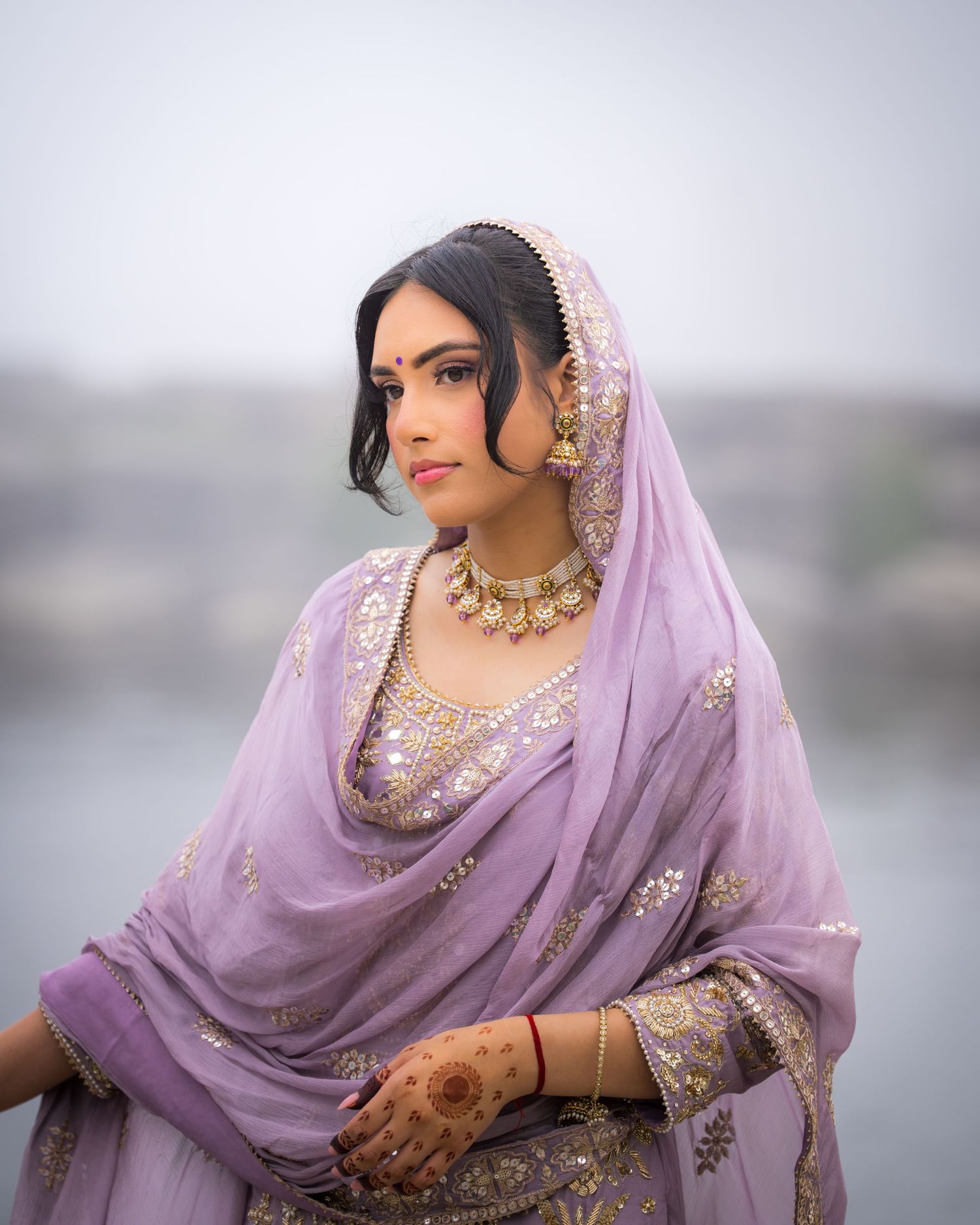 A person in a lavender embellished outfit with jewelry poses elegantly against a misty backdrop.
