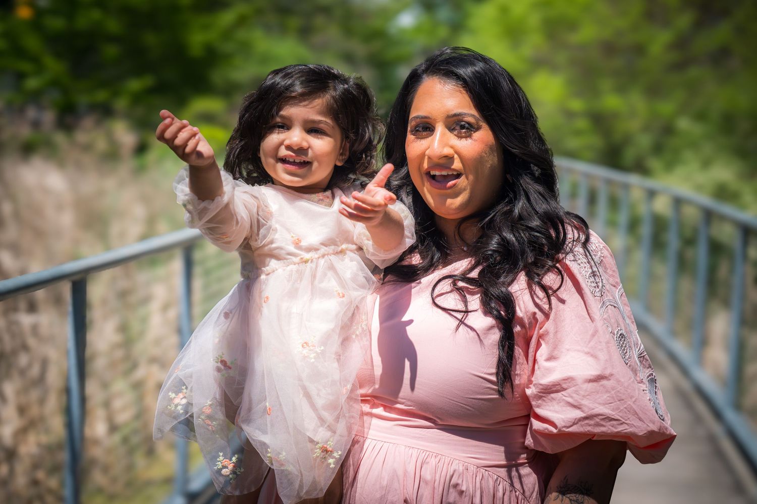 A woman in a pink dress holds a toddler girl in a white dress on a sunny bridge outdoors.