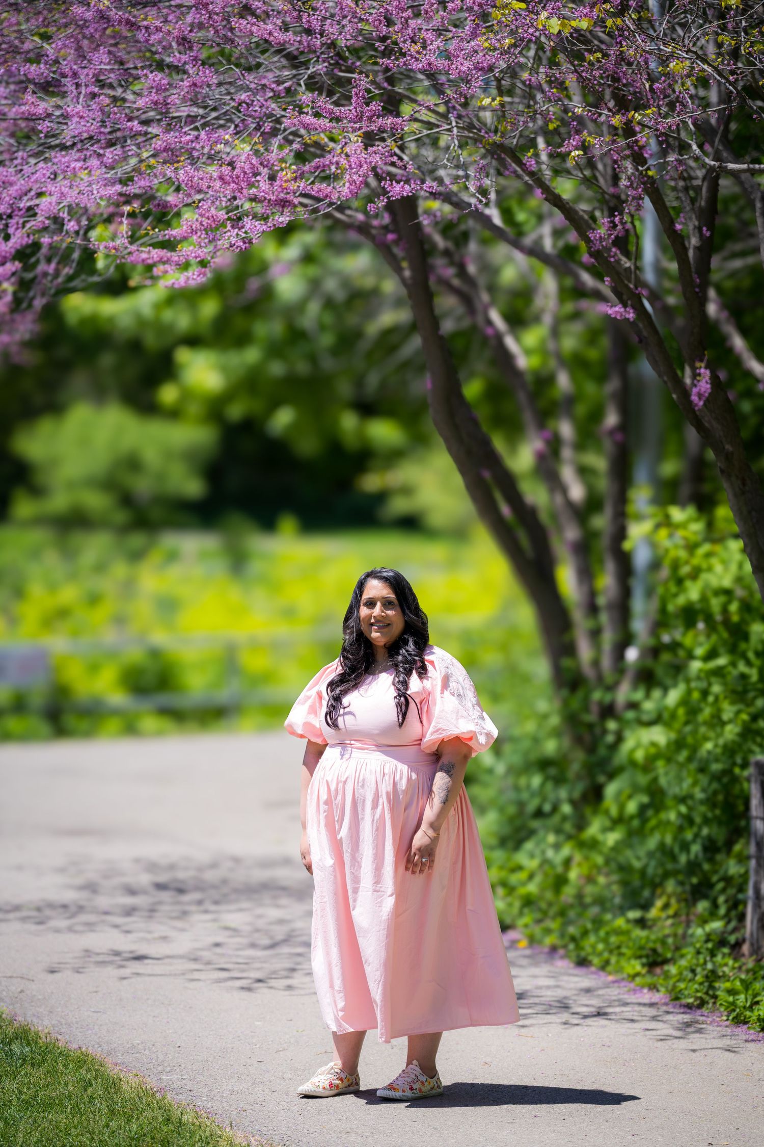 A pregnant woman in a pink dress walks under blooming purple redbud trees on a sunny path.
