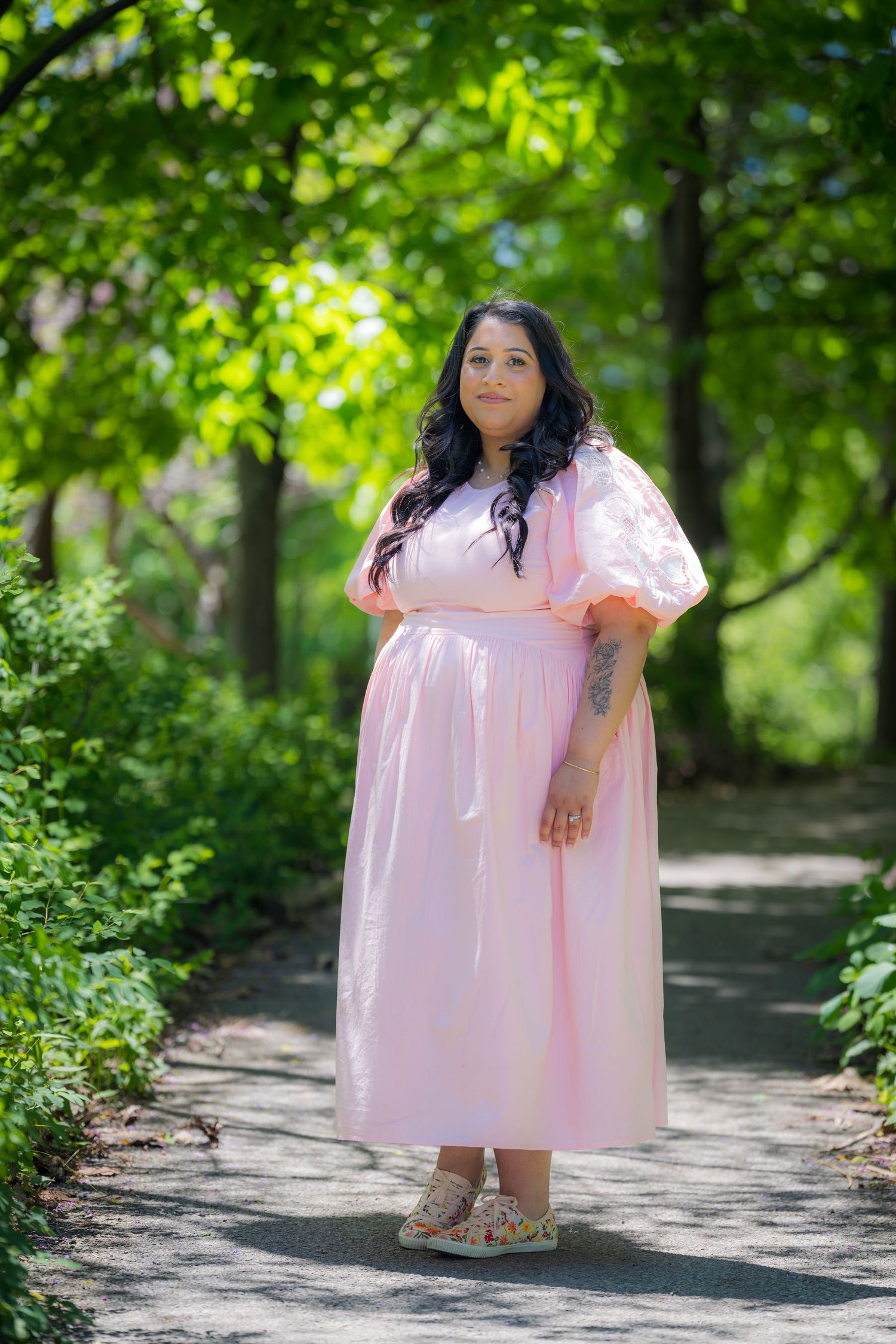 Woman in flowing pink midi dress standing on garden path surrounded by lush green trees.