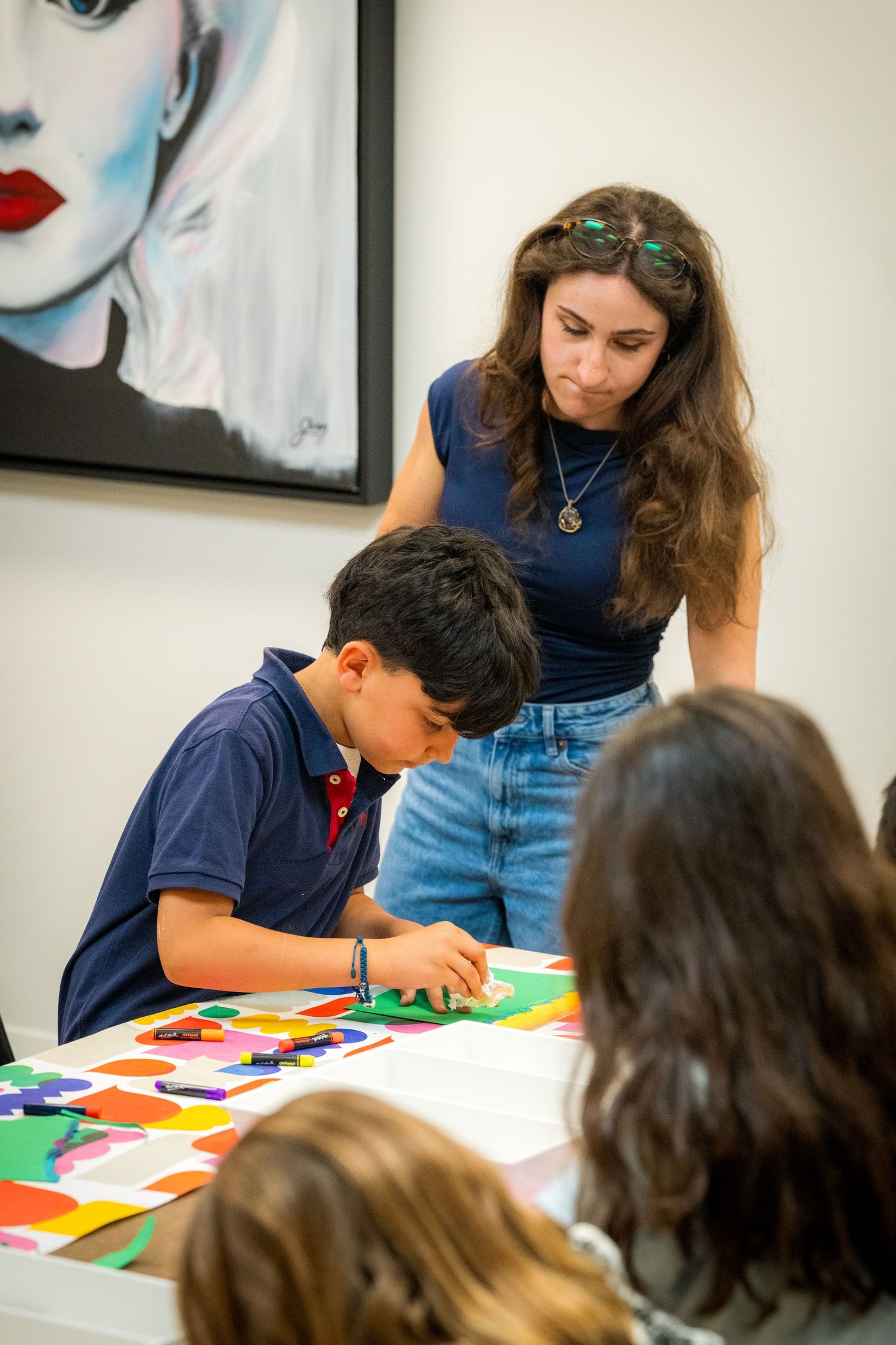 A female art instructor supervises a young boy creating a vibrant paper collage in an art gallery.