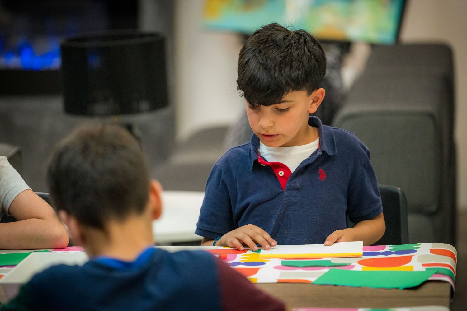 A focused young boy works on a colorful art project at a table with craft supplies.