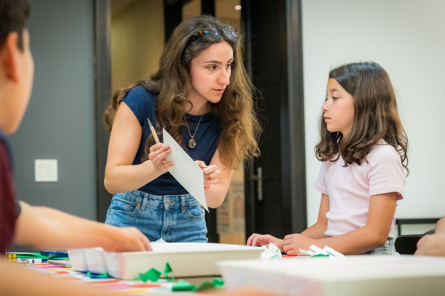 Teacher guides young girl in origami craft activity at colorful art table in classroom setting.