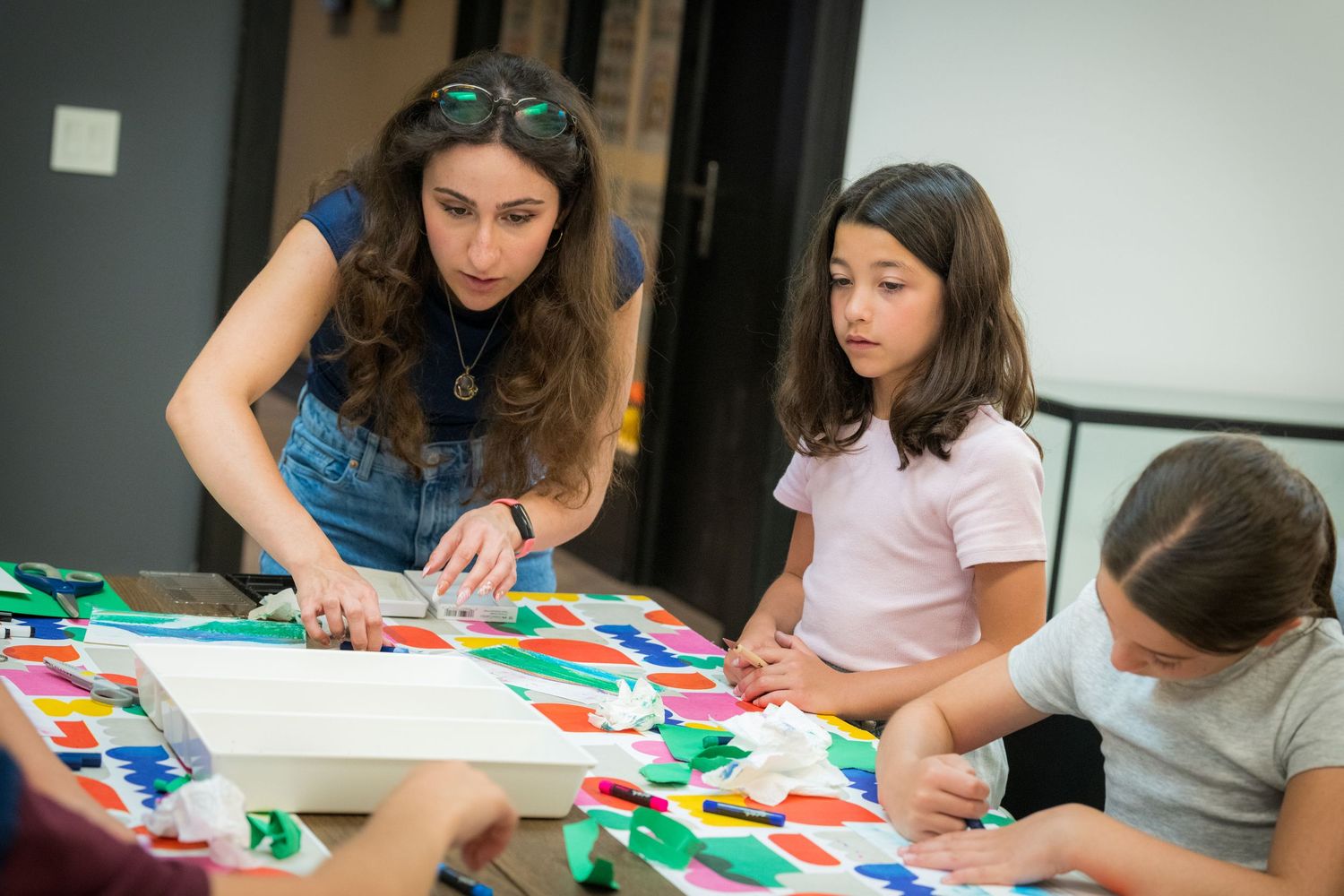 A female teacher guides two young girls in creating a colorful paper collage artwork.