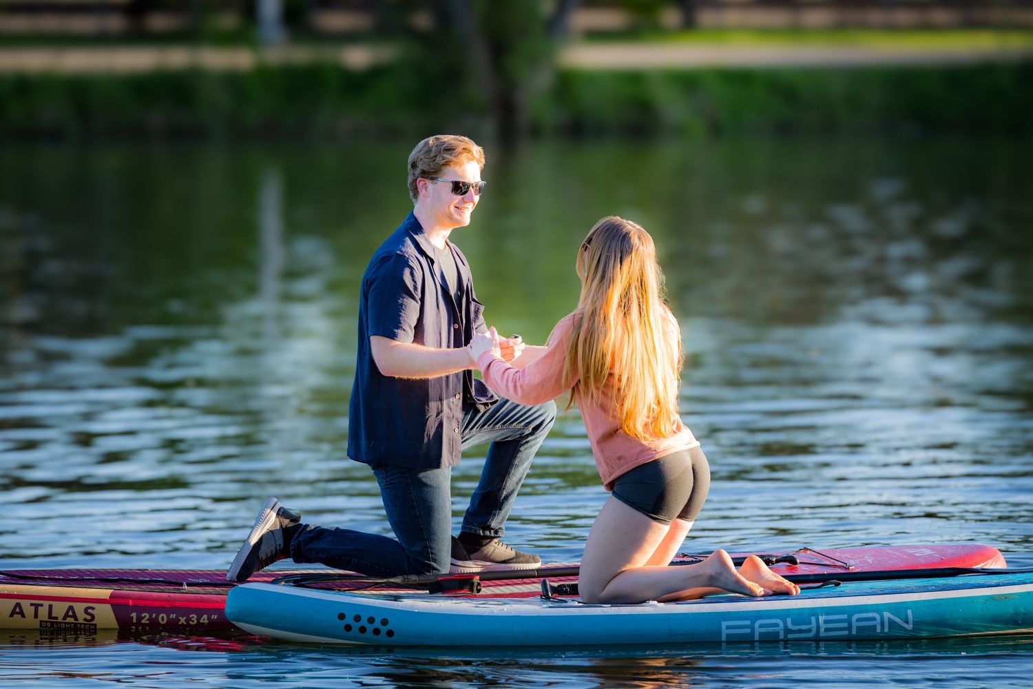 Man kneeling on paddleboard proposing to woman on a calm sunny lake.